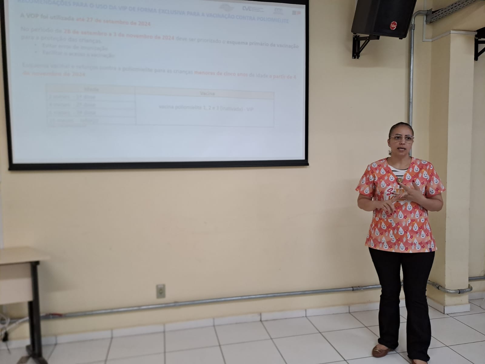 Imagem mostra um telão ao fundo com diversas informações escritas em preto com fundo branco e a palestrante a frente, uma mulher de cabelos comprido amarrado