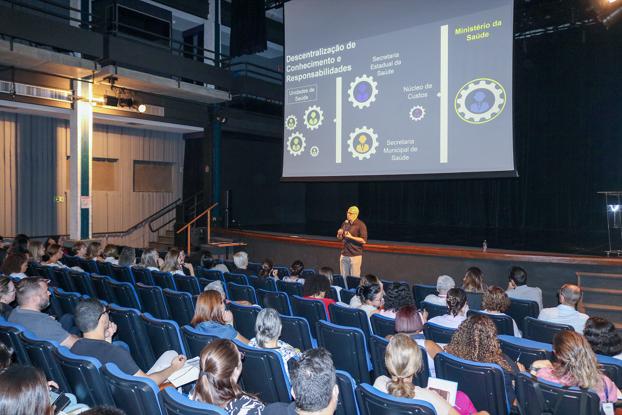 imagem mostra a sala do teatro repleta de pessoas assistindo a uma palestra. ao fundo um homem fala para o público em frente de um telão com informações de saúde