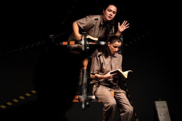 Duas mulheres, vestidas com macacão bege estão numa estrutura. Uma sentada, lendo um livro e outra de pé atrás dela e acima, com um livro na mão e a outra mãoe espalmada para frente. O fundo da imagem é preto.