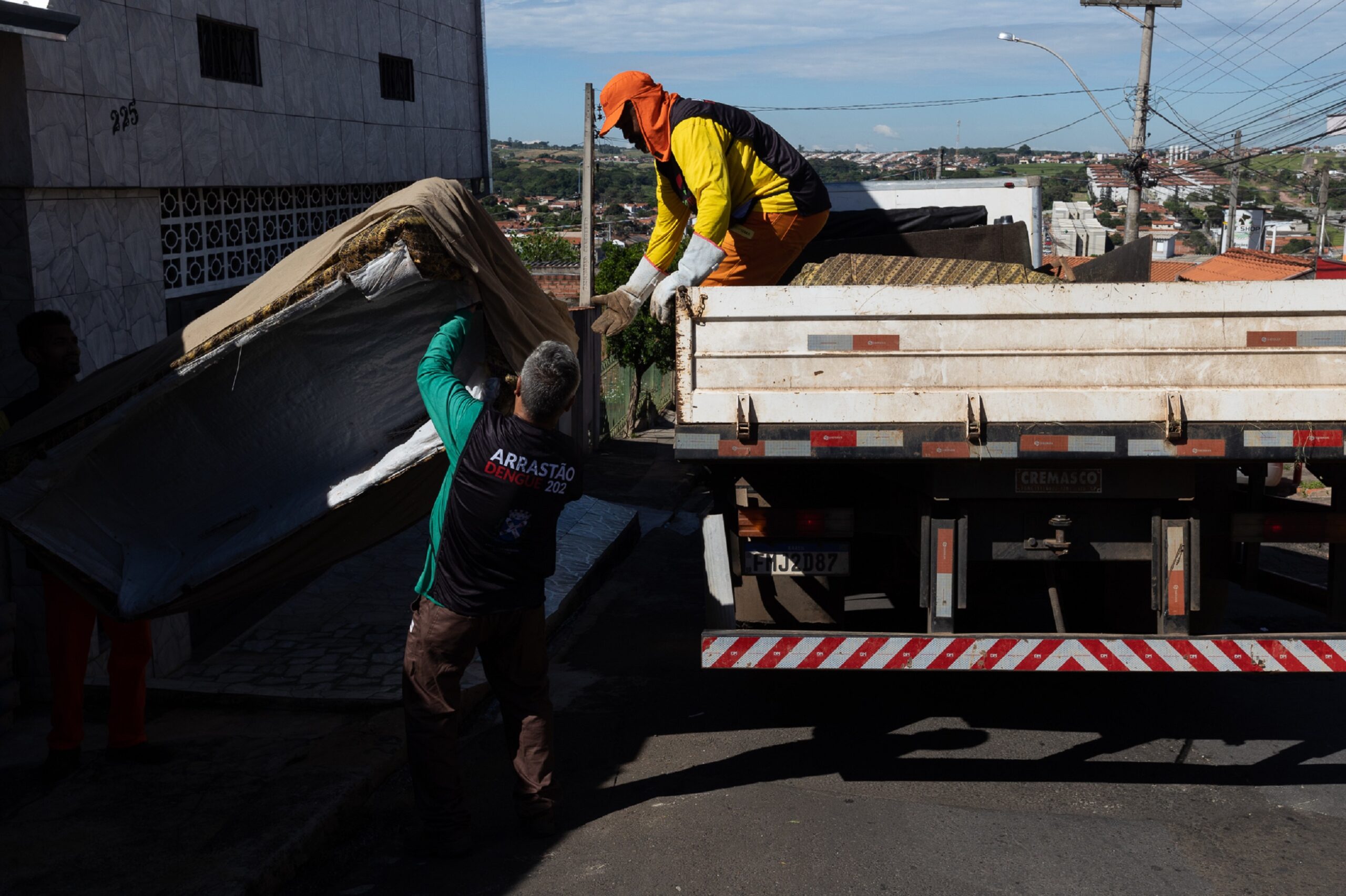 Imagem mostra a ação de dois trabalhadores fazendo o recolhimento de inservíveis na rua. um está sobre a carroceria de uma caminhão de colete amarelo e outro está no chão com colete azul subindo uma grande mesa velha de madeira, o dia está ensolarado