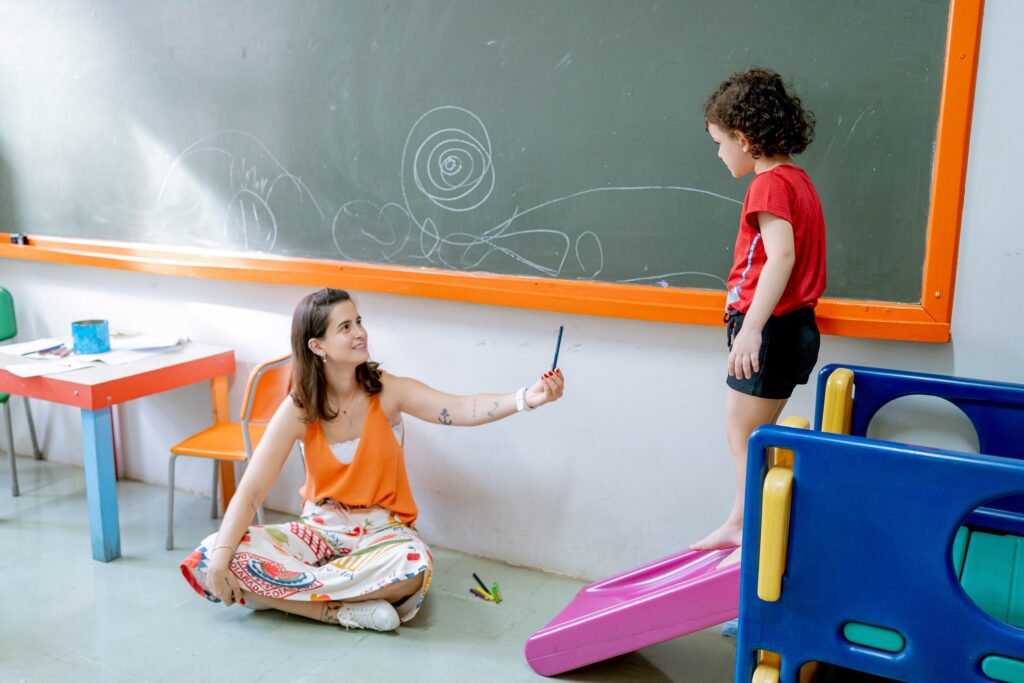Na imagem, em uma sala de aula, uma professora está sentada no chão com as pernas cruzadas e oferece uma caneta para uma menina realizar suas atividades expostas na louse verde ao fundo