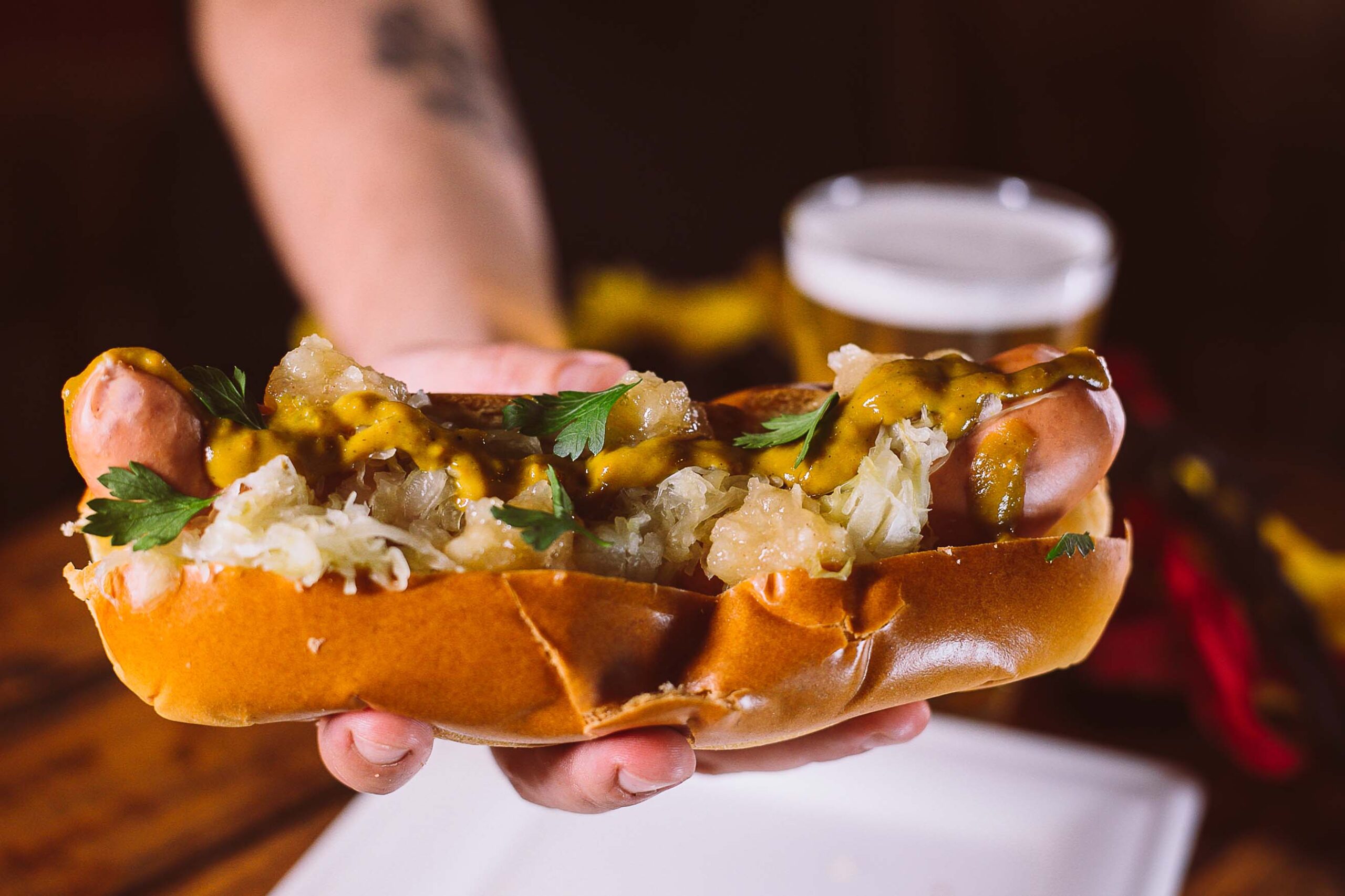 Descrição da Imagem: Um close-up de um cachorro-quente sendo segurado por uma pessoa. O cachorro-quente está em um pão macio e é coberto com chucrute, mostarda e salsa. Um copo de cerveja desfocado está ao fundo. A pessoa que segura o cachorro-quente tem uma tatuagem no braço.