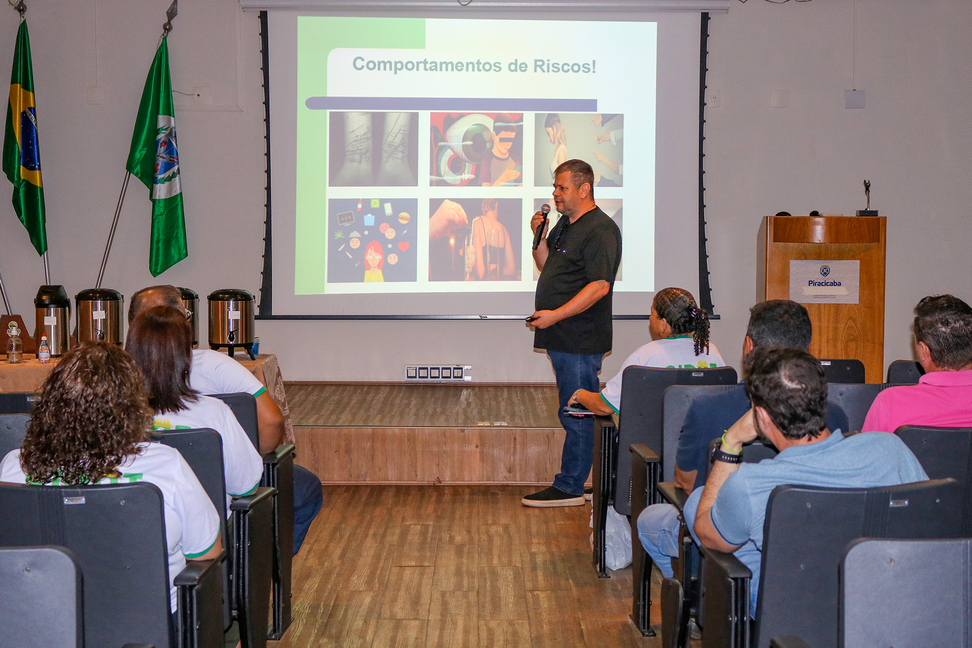 Imagem mostra um grande anfiteatro, repleto de pessoas sentadas em cadeiras assistindo a uma palestra ministrada por um psicólogo homem, estatura média, cabelos brancos, usando calça jeans e camiseta preta, ao fundo um telão reproduzindo imagens alusivas ao tema da palestra que é ISTs