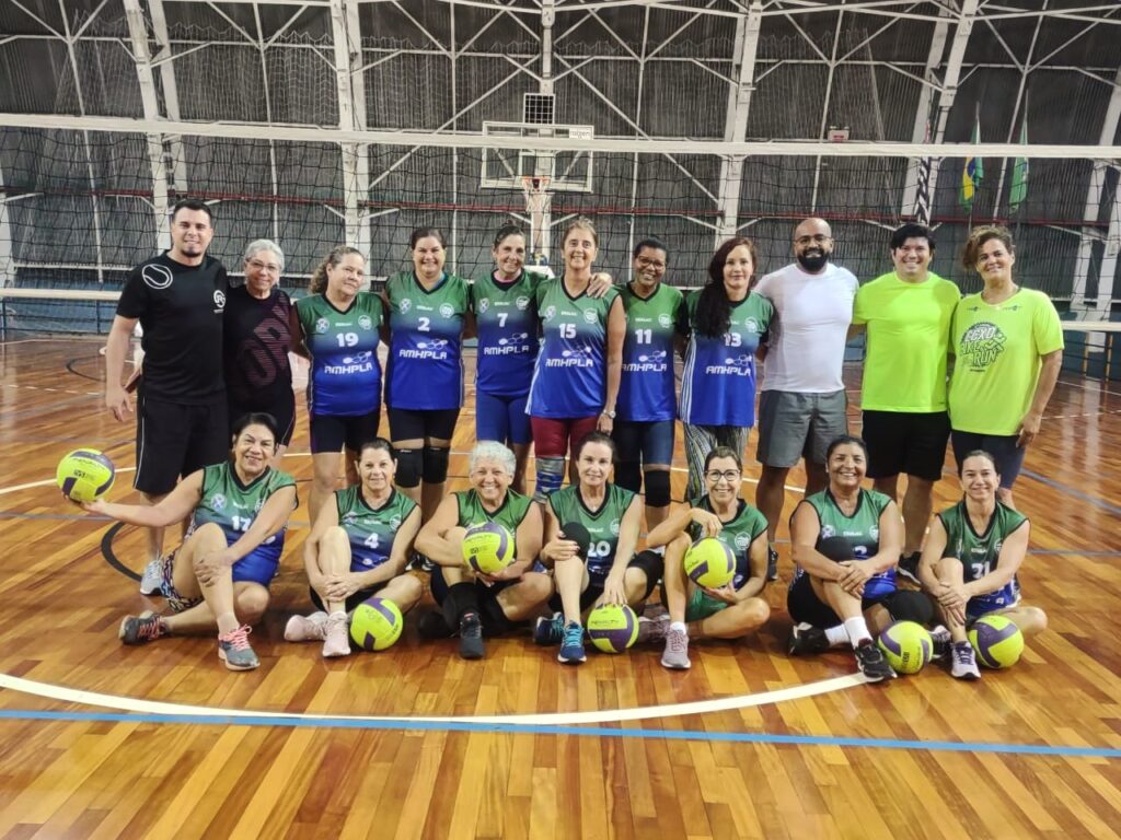 Na imagem aparece a equipe de voleibol adaptado feminino. As atletas estão com uniforme verde, acompanhadas dos técnicos e de outras pessoas, vestindo roupas nas cores preta, branca e amarela.