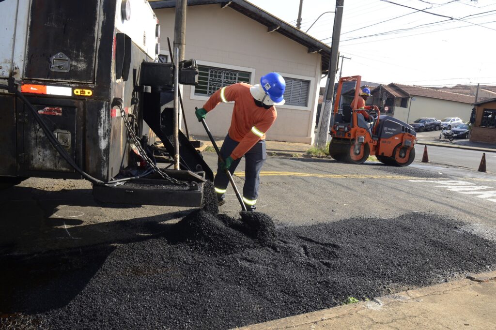 Homem com capacete azul e uniforme com camisa laranja e detalhes verde claro e branco e calça cinza, mexe com uma pá na massa asfáltica.