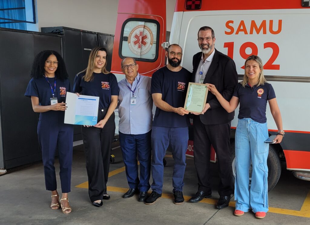 imagem mostra uma ambulância do Samu 192 ao fundo e, na frente, seis pessoas da secretaria da saúde segurando certificado de registro no CRM e CFM e a nova portaria do NEU