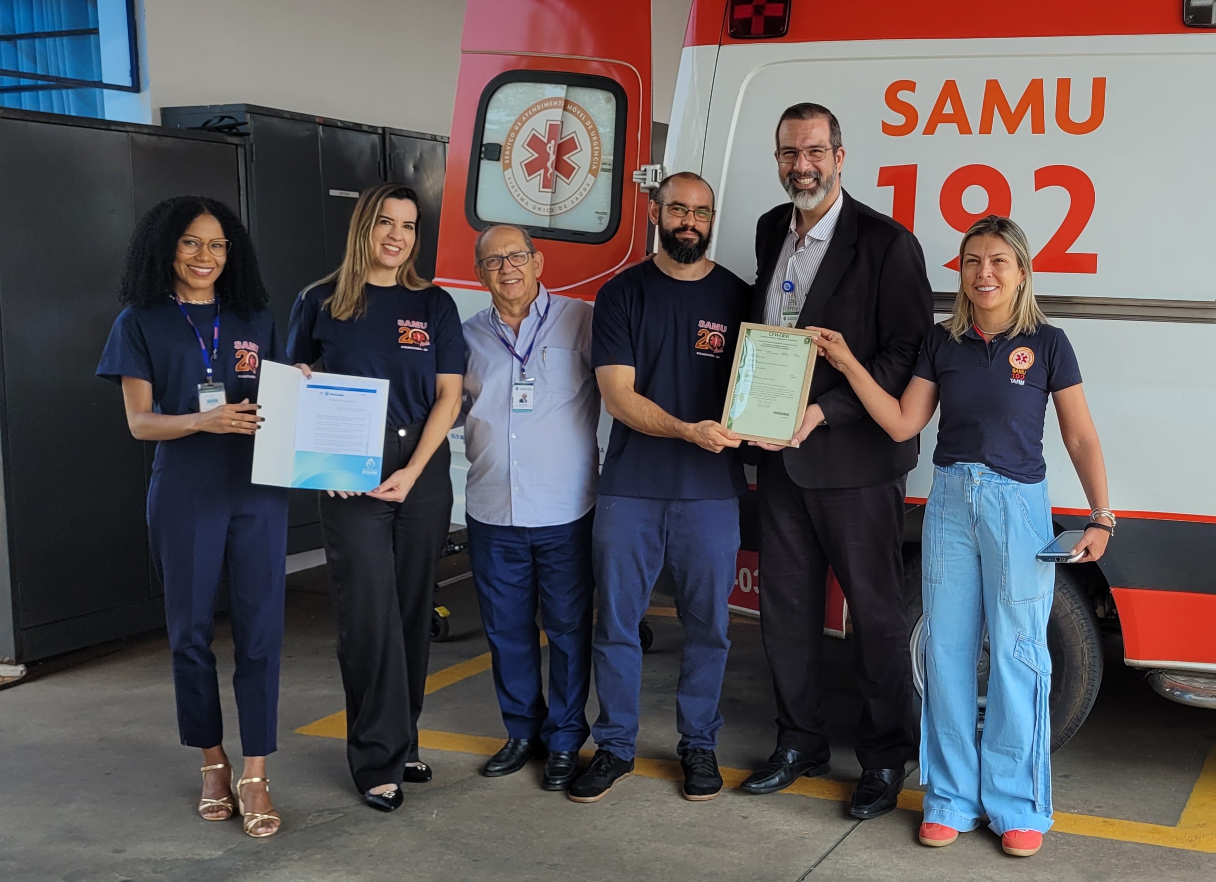 imagem mostra uma ambulância do Samu 192 ao fundo e, na frente, seis pessoas da secretaria da saúde segurando certificado de registro no CRM e CFM e a nova portaria do NEU