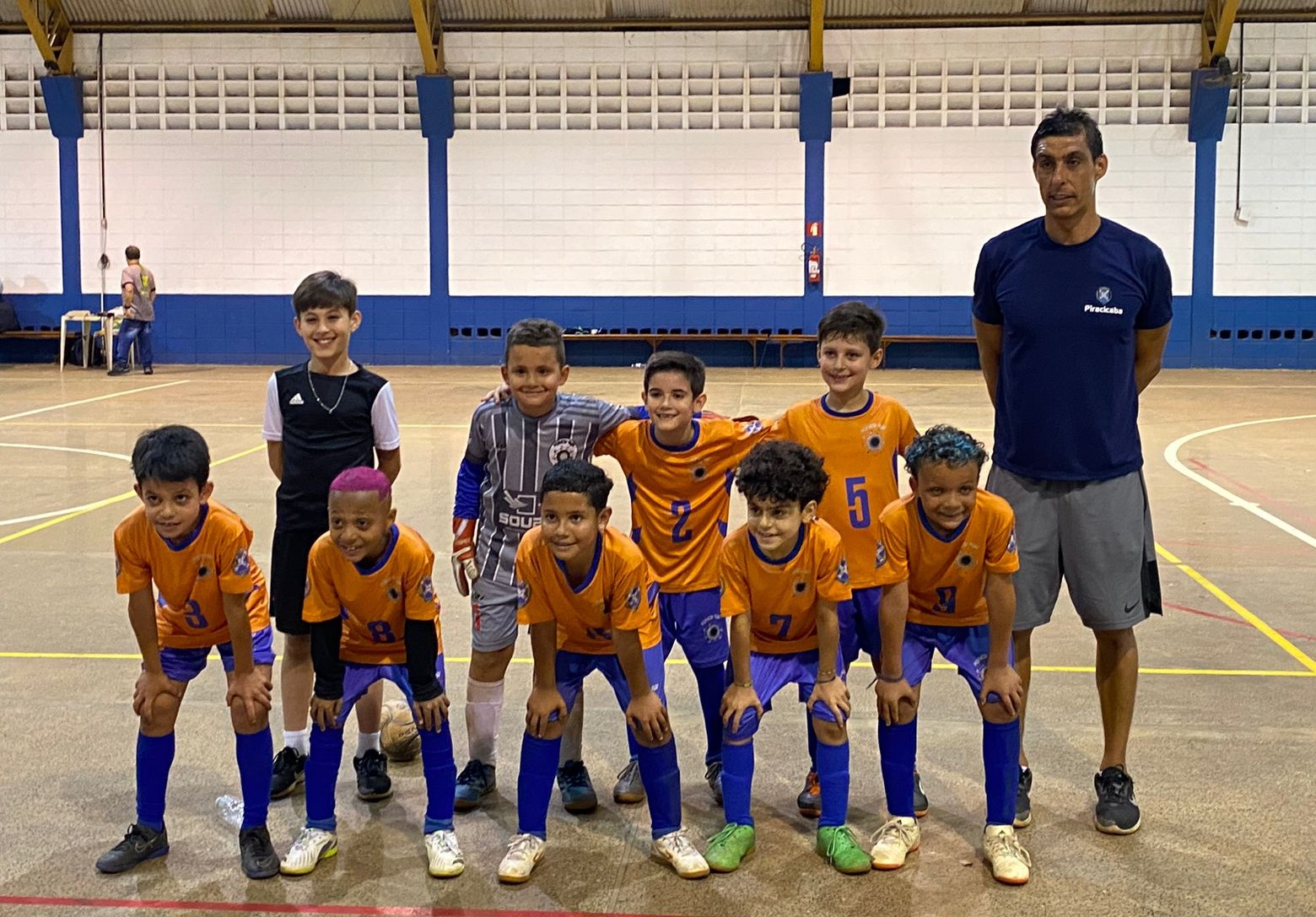 Equipe de futsal com crianças da categoria sub-8 de Piracicaba estão olhando para a produção de uma fotografia. Elas estão com uniforme nas cores laranja para a blusa e, azul, para o calção. O técnico está vestindo uma camisa azul com short cinza. Eles estão posando para a foto num ginásio com paredes brancas e com faixas azuis em destaque.