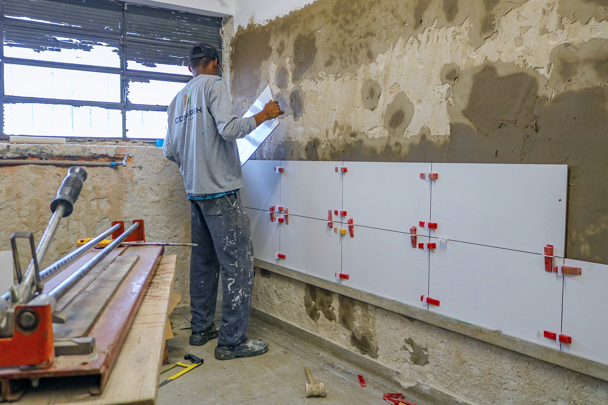 Na imagem, um homem está segurando uma peça de cerâmica quadrada na cor branca e faz a sua fixação na parede; o homem já fez a colocação de 12 dessas peças cerâmicas