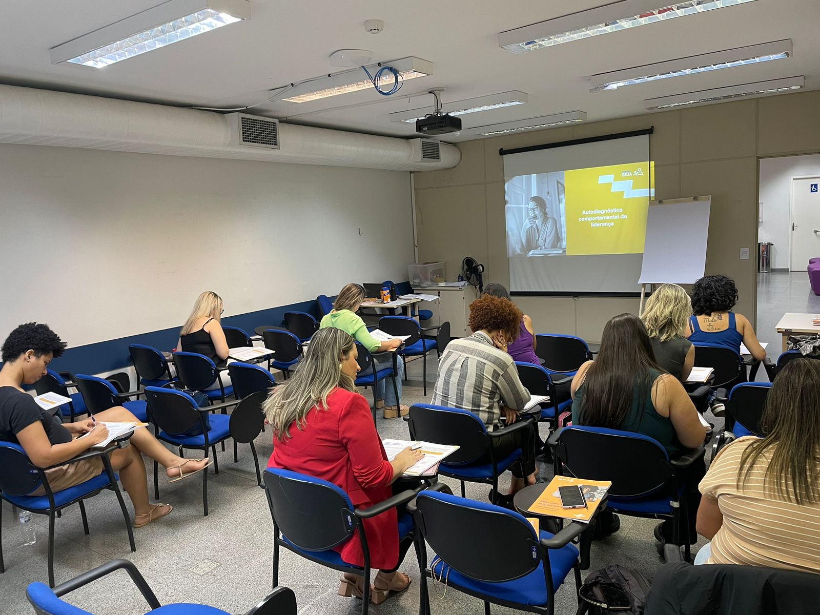 Imagem mostra uma sala de aula repleta de mulheres sentadas em carteiras fazendo atividade coletiva sobre assunto empreendedorismo. ao centro um telão exibe a atividade realizada pelas mulheres