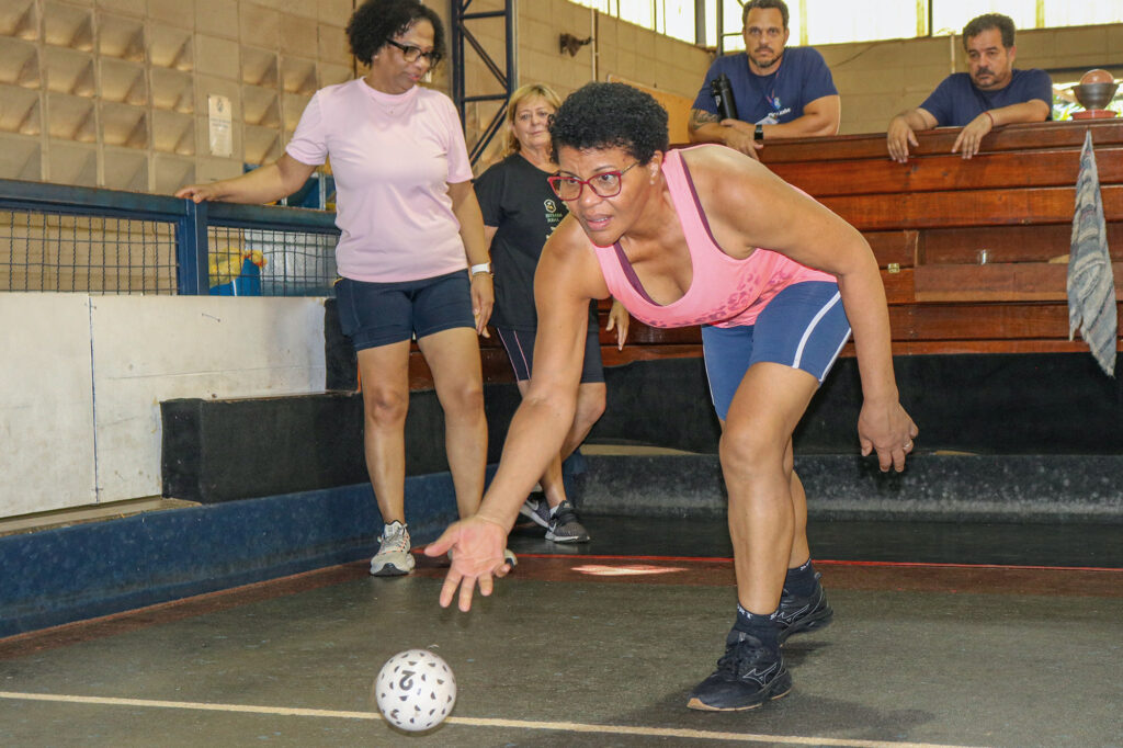 Marlene dos Santos de Camargo, de 58 anos, arremessa a bola de bocha.Ela está vestindo camiseta rosa com calção azul. A bola de bocha é branca com detalhes em preto.Ao fundo, quatro pessoas acompanham o arremesso de Marlene.
