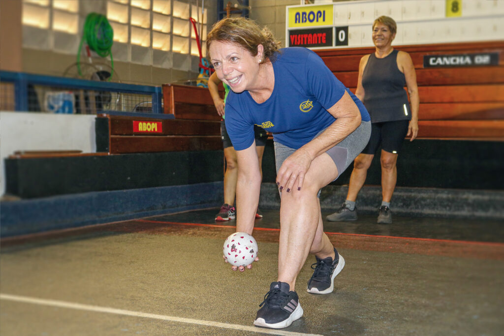Nilce Tobaldini, de 64 anos, sorri enquanto arremessa a bola da bocha.Ela veste roupa azul com calção cinza. Sua companheira de equipe está logo atrás, observando sua jogada.