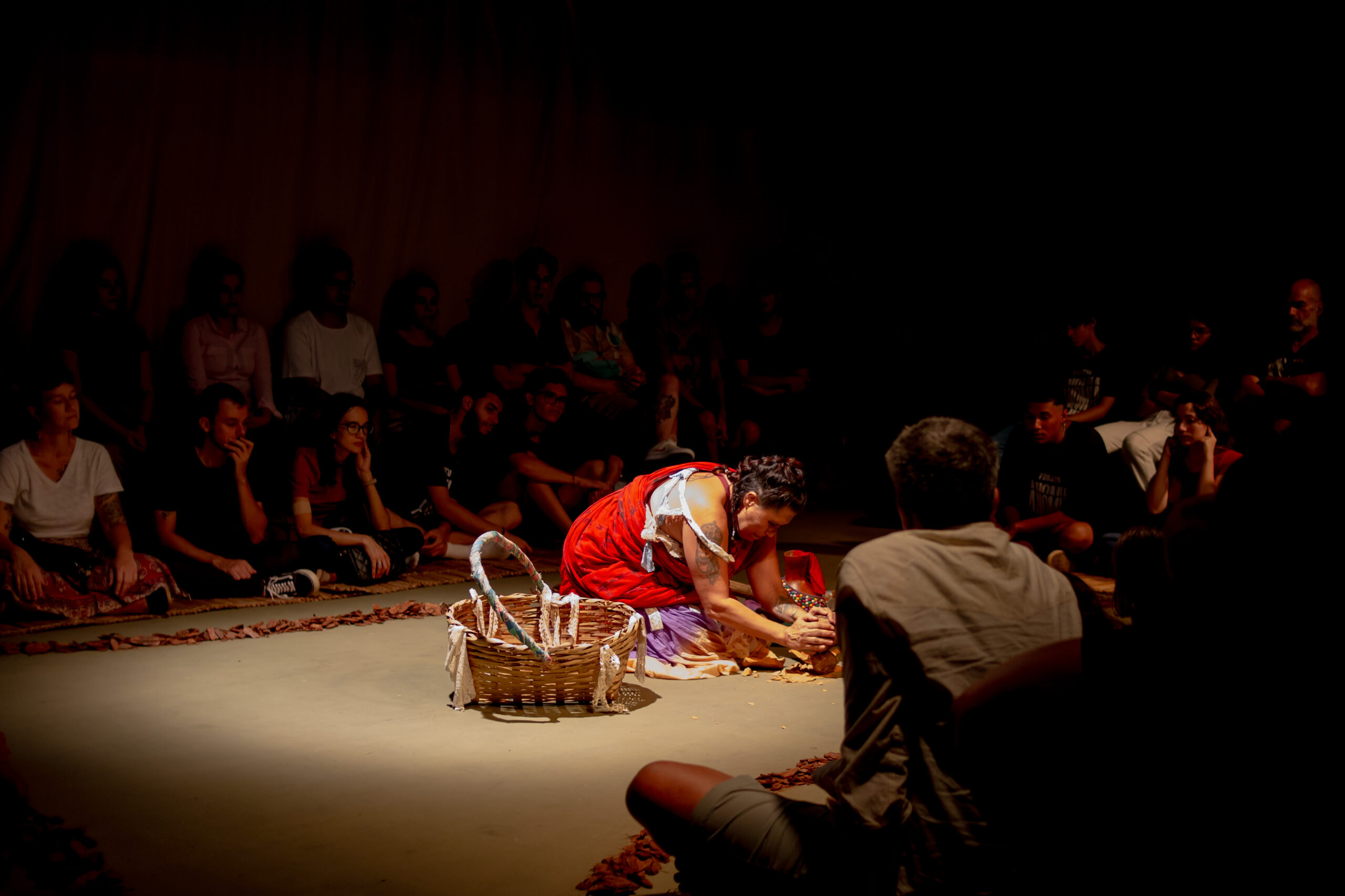 Na imagem, cena do espetáculo vai-o-vento. com a luz centralizada em um círculo, a atriz agachada, estende suas mãos.