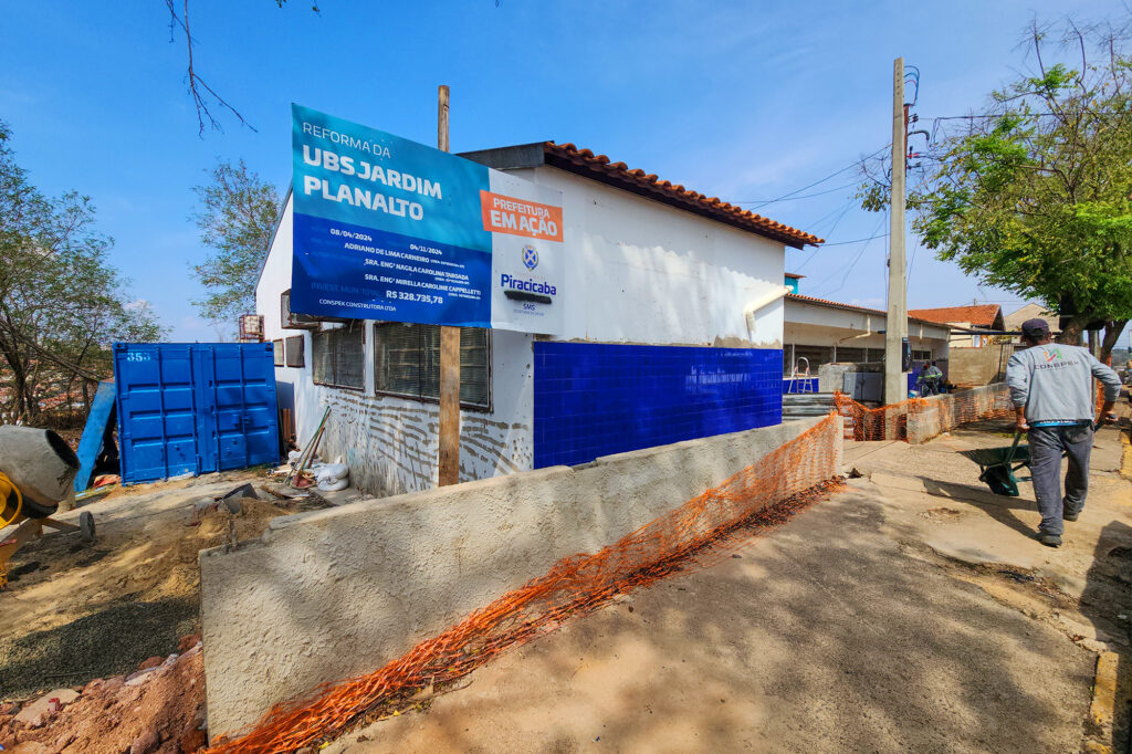 Imagem mostra a frente da unidade em obras. Da esquerda para a direita vemos a construção de um muro de tijolo e concreto, a unidade está pintada de branco com cerâmica azul já instalada na cor azul até um metro de altura, à direita um trabalhador empurra um carrinho de pedreiro com materiais que serão utilizados no interior da obra
