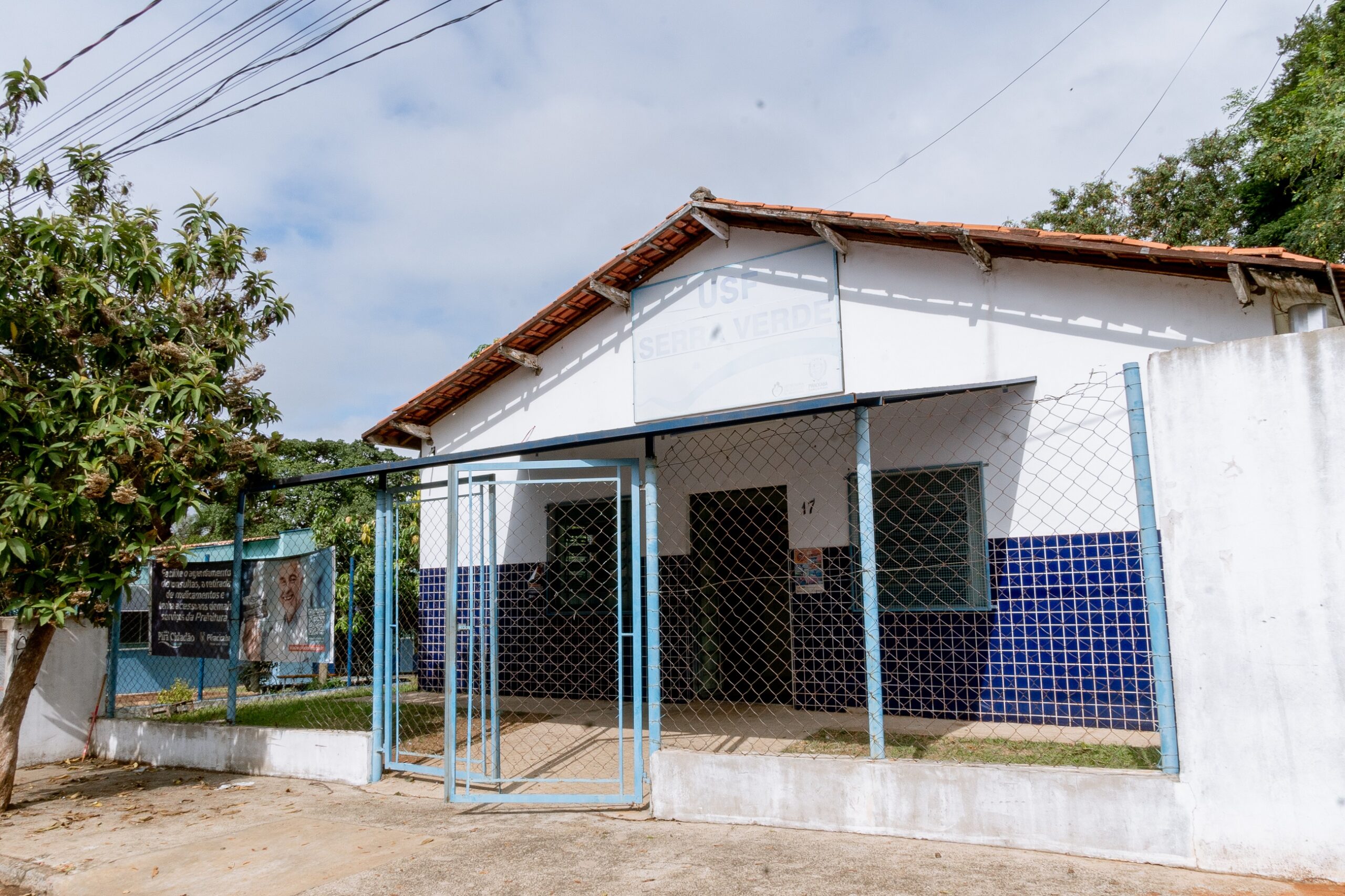Imagem mostra a fachada da unidade nas cores azul e branca desbotado pelo tempo e com o portão de ferro, azul, aberto para entrada da população