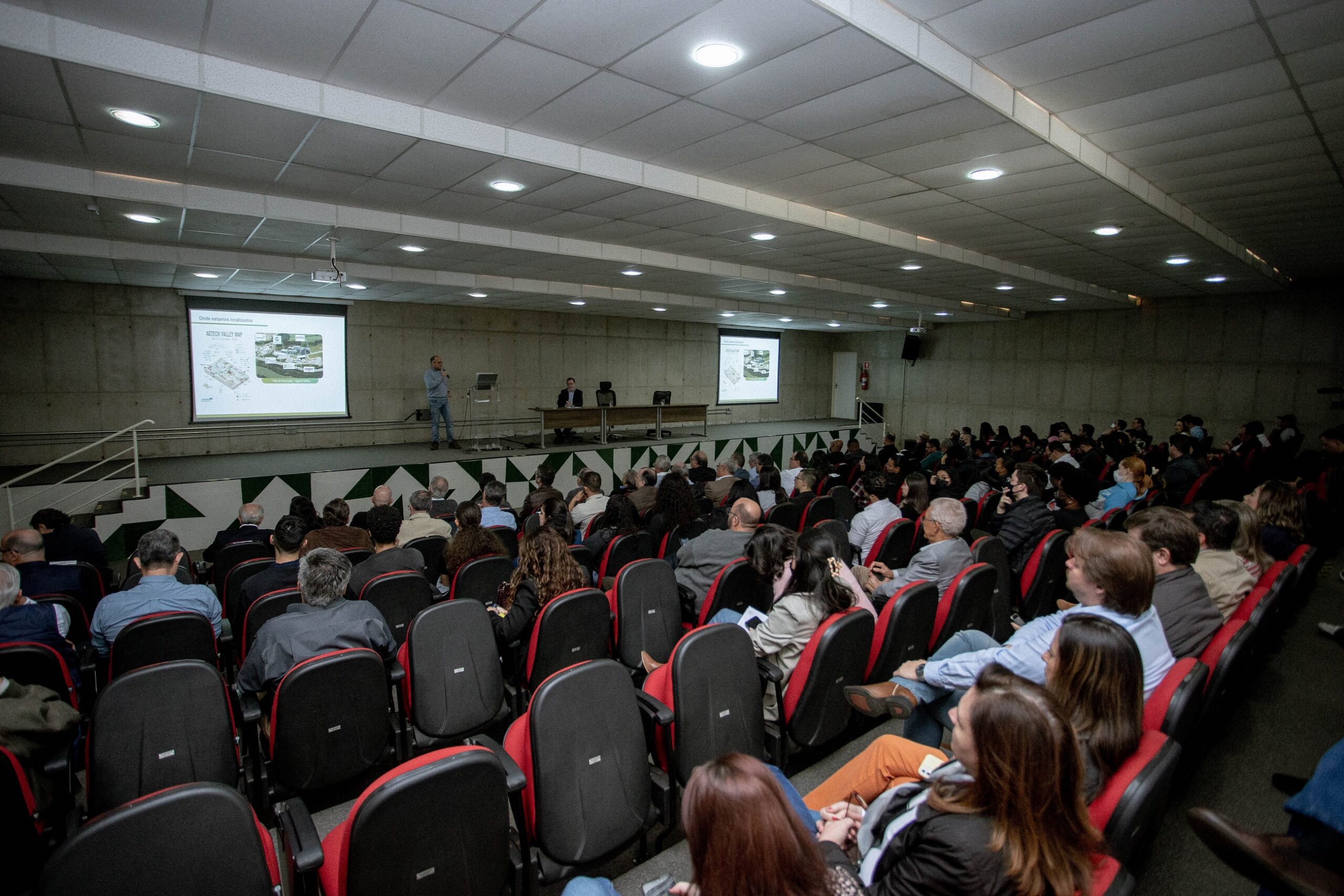 imagem mostra um auditório com mais de 100 cadeiras, todas ocupadas por pessoas; a frente existe um telão com informações sobre a palestra enquanto um homem de terno e gravata fala para as pessoas