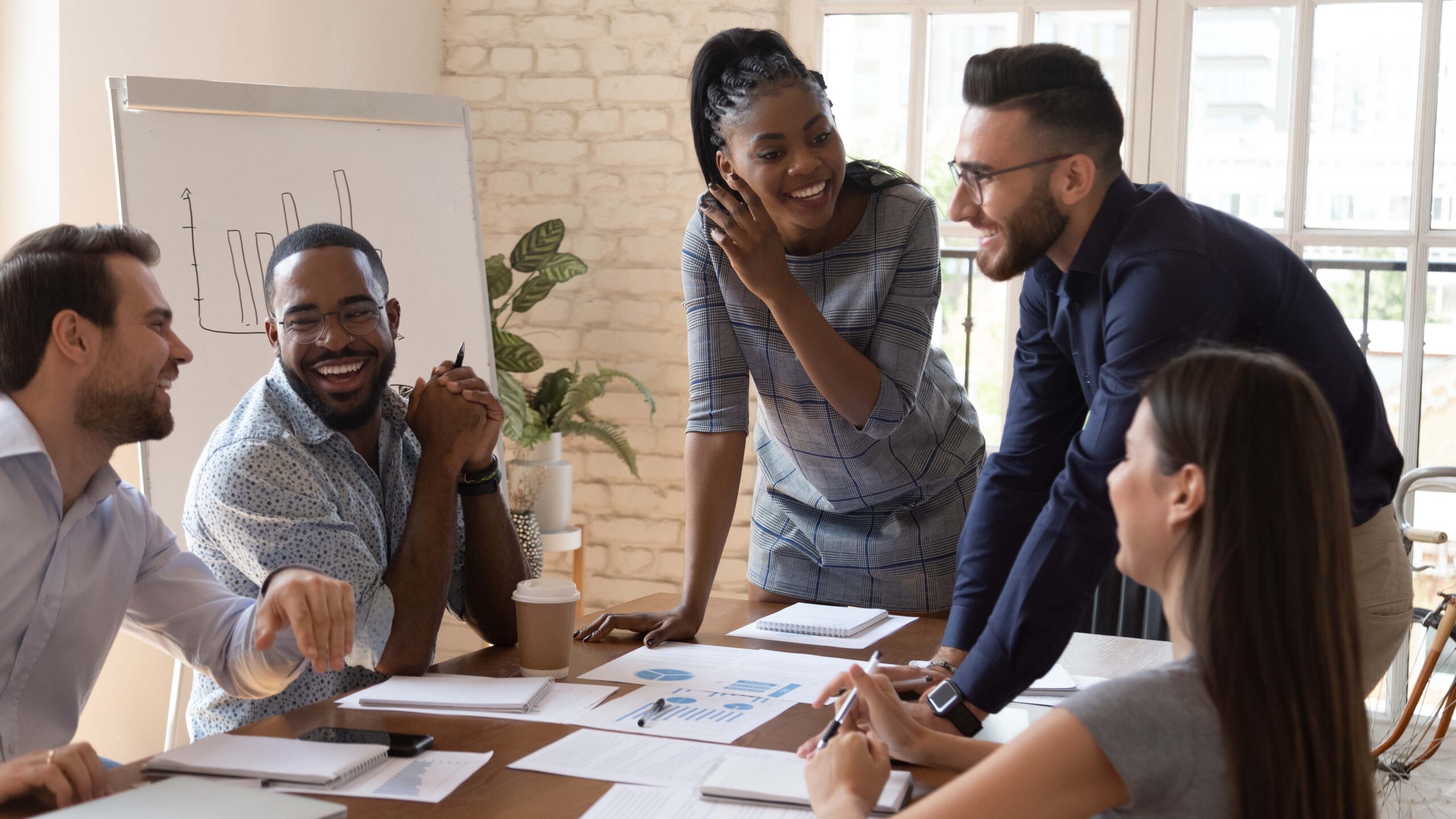 em uma mesa grande de madeira, cinco pessoas trabalham na organização interna da empresa em meio a uma reunião com diversos papeis na mesa; um homem e uma mulher estão em pé sorrindo e dois homens e uma mulher estão sentados ouvindo o que eles dizem