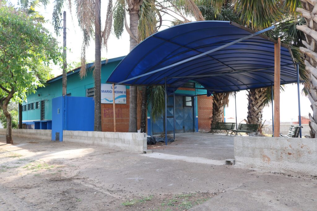 Imagem mostra a frente da unidade Mário Dedini quase com as obras concluídas. à direita mostra a nova entrada com cobertura em estrutura de ferro e telhado de acrílico, ao redor da unidade já está toda pintada nas cores azul e verde escuro