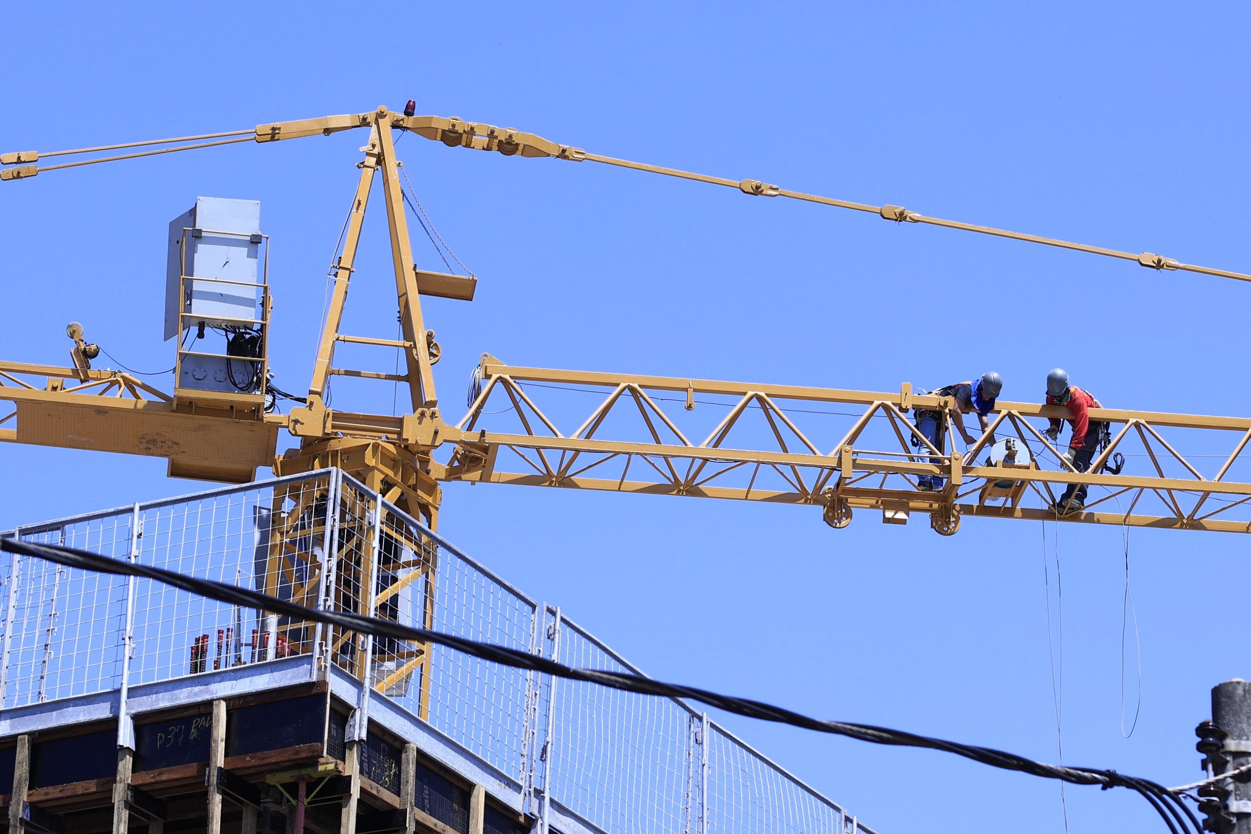 Imagem mostra um céu azul distante e, bem no alto, um guincho de construção civil feito para edificar prédios com dois homens andando por cima deles usando cintos e cordas como equipamentos de segurança, alem de capacete e luvas
