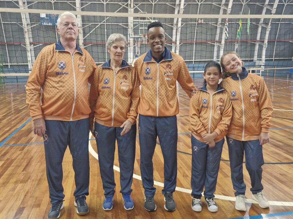 Cinco atletas, de várias gerações, estão sorrindo durante produção de fotografia na apresentação dos novos uniformes que serão usados em competições oficiais.Os uniformes foram produzidos na cor laranja e com calção na cor azul.