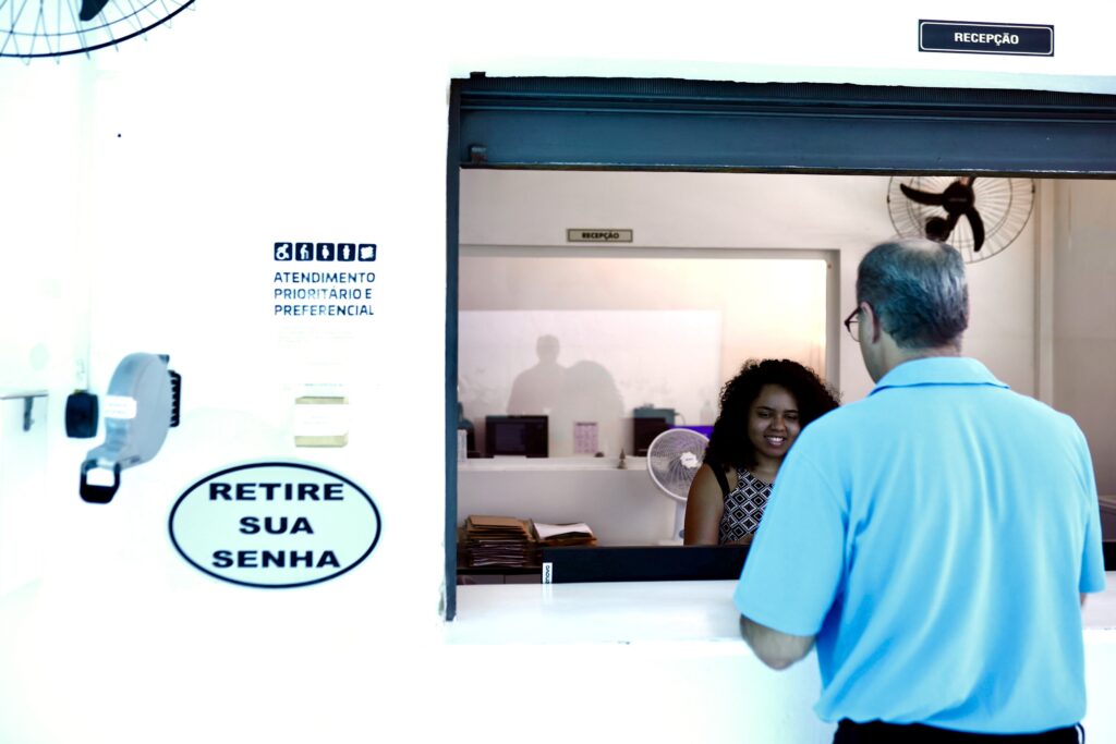 A imagem mostra uma recepção. Há uma mulher jovem com cabelo escuro sentada à espera de atendimento. Um homem de meia-idade, com cabelo curto e grisalho e óculos, de costas para a câmera, está a aproximar-se do balcão. Há um sinal na parede que diz "Retire sua senha" e outro que indica atendimento prioritário e preferencial. Há também um dispensador de senhas. O ambiente parece ser um local de atendimento ao público.