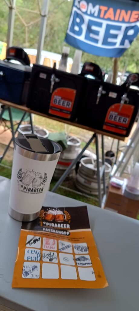 A imagem mostra um copo de cerveja personalizado com a logomarca "Pirabeer" em cima de uma mesa. Ao fundo, vêem-se vários recipientes de cerveja, provavelmente barris ou recipientes para transporte, e uma faixa ou bandeira com a inscrição "Komtaine Beer". Há também um cartão ou caderneta com vários espaços para carimbos, indicando que possivelmente é um evento de degustação ou festival de cerveja, onde o participante deve carimbar seu cartão ao provar diferentes cervejas. A cena sugere um ambiente de festival ao ar livre ou de uma feira de cerveja. O copo de cerveja está próximo ao cartão, sugerindo que é para um participante do evento.