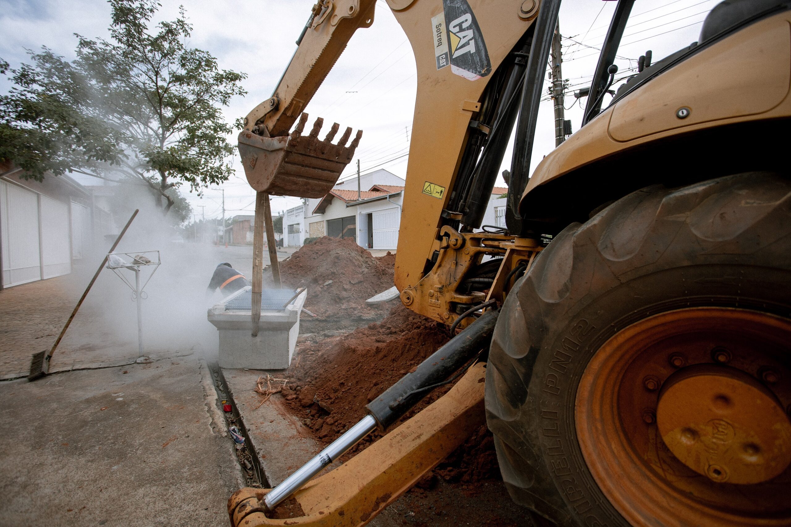 A imagem mostra uma escavadeira hidráulica amarela, da marca Caterpillar (CAT), trabalhando em uma rua. A escavadeira está parcialmente enterrada no chão, com sua caçamba voltada para baixo. Há um monte de terra recém-escavada ao lado dela. No fundo, é possível ver casas e árvores, indicando um ambiente residencial ou urbano. Há uma pessoa quase invisível, encoberta por uma fumaça ou vapor, perto de um bueiro de concreto. A cena sugere que estão realizando obras de infraestrutura na rua, como a instalação ou reparo de algum tipo de encanamento ou rede subterrânea. A fumaça ou vapor pode indicar a utilização de algum tipo de equipamento ou produto que gera essa liberação. A imagem está um pouco escura e a atmosfera geral é de trabalho em progresso em uma área residencial.