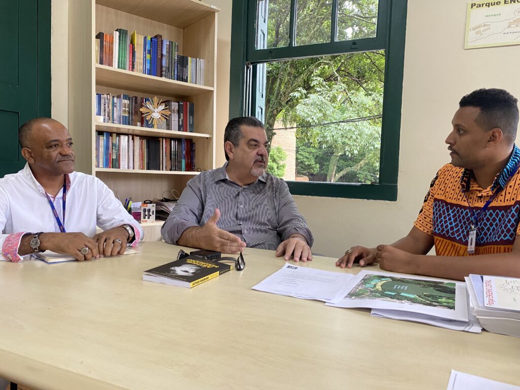 Três homens estão sentados de um lado de uma mesa, conversando. Na mesa, aparecem papeis.