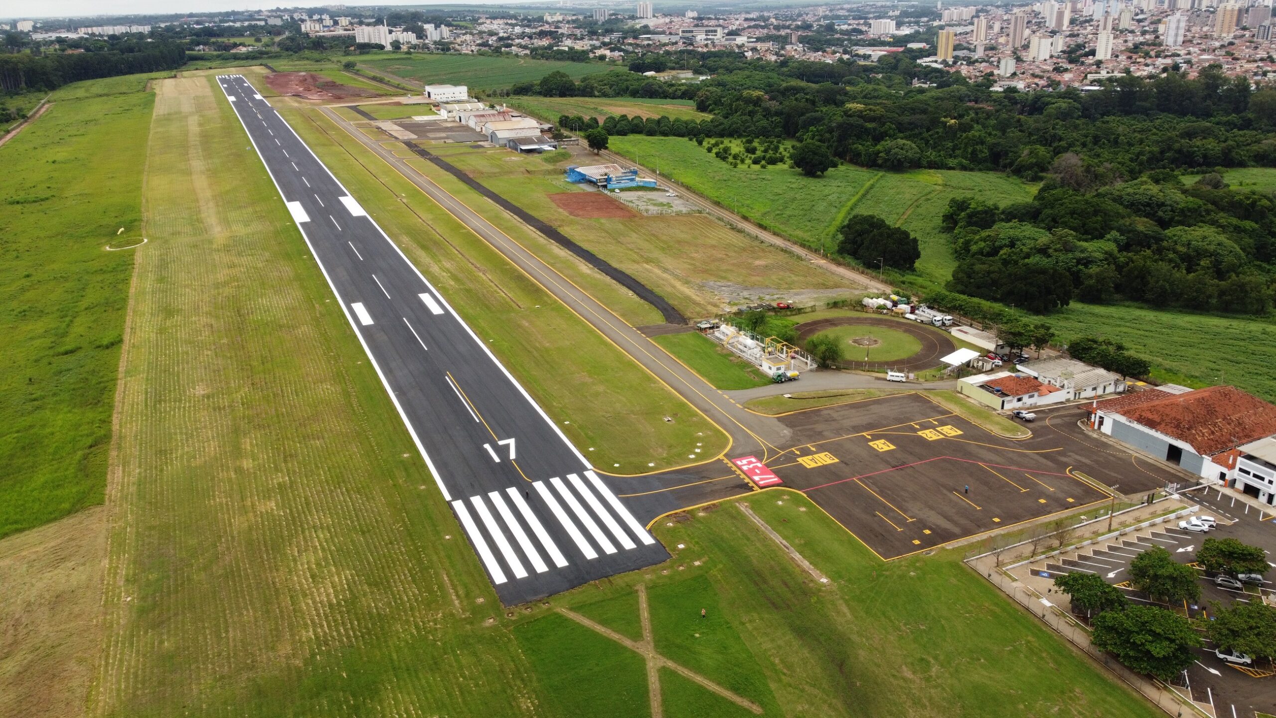 A imagem é uma vista aérea de uma pista de pouso de aeroporto, mostrando a pista principal, a área de taxiamento e as instalações do aeroporto. A pista de pouso é longa e reta, com marcações brancas bem definidas. A numeração "17" está visível na pista, indicando a sua orientação. A área de taxiamento é escura e apresenta marcações amarelas que indicam as posições de estacionamento de aeronaves. Há também um heliponto circular próximo às instalações do aeroporto. As instalações incluem um hangar de tamanho considerável e vários edifícios menores. O entorno do aeroporto é composto principalmente por áreas verdes, com vegetação abundante e áreas cultivadas. Ao fundo, é visível uma cidade com prédios de diferentes tamanhos e alturas. A imagem sugere um dia ensolarado com boa visibilidade. A imagem demonstra a infraestrutura de um aeroporto relativamente pequeno, mas bem organizado e integrado à paisagem ao seu redor.