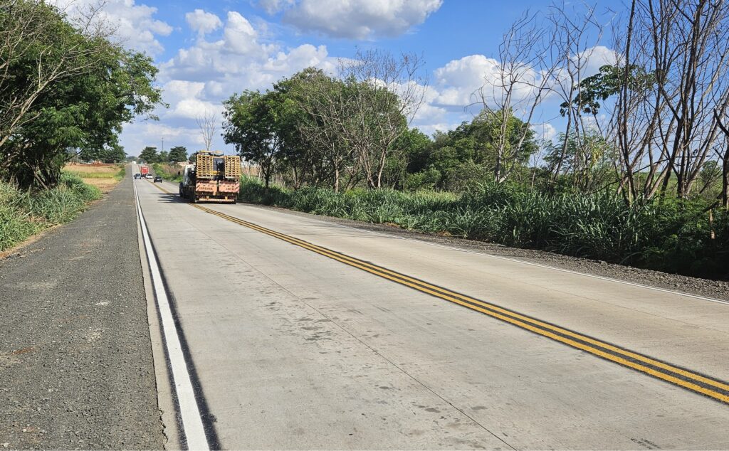 Imagem da rodovia já pavimentada com concreto e sinalizada; há árvores dos dois lados e, ao fundo, um caminhão e alguns carros passando