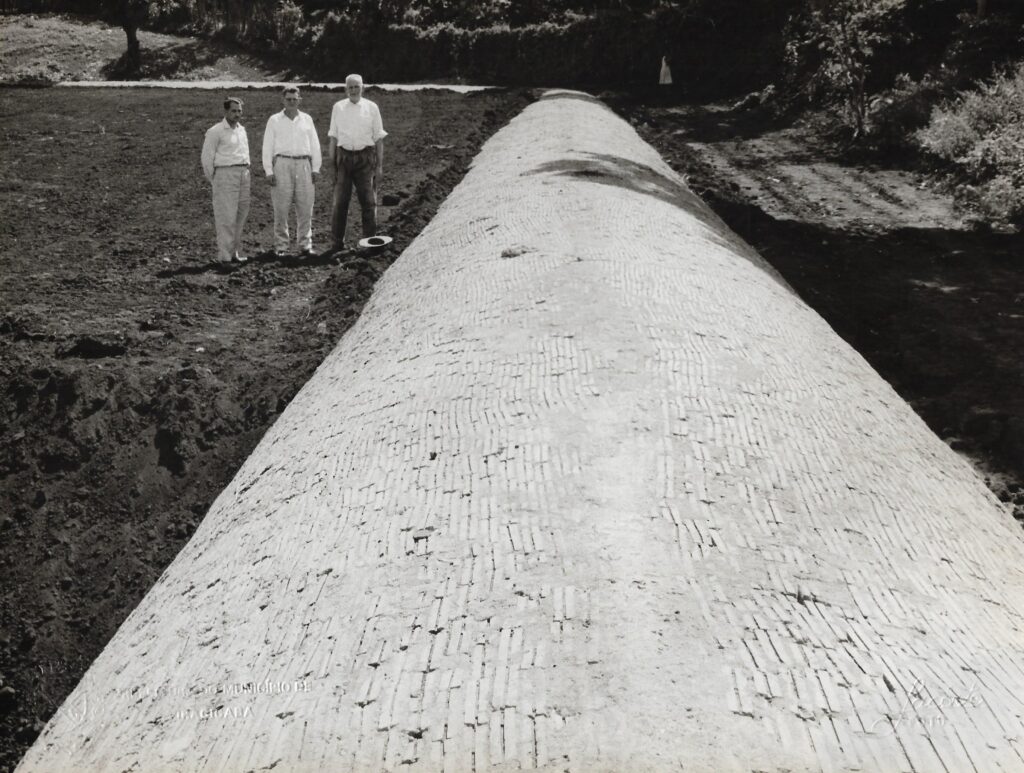 Imagem antiga, mostrando a canalização recém concluída; do lado esquerdo, há três homens em pé, dois vestindo camisas brancas e calças claras e um com camisa branca e calça escura