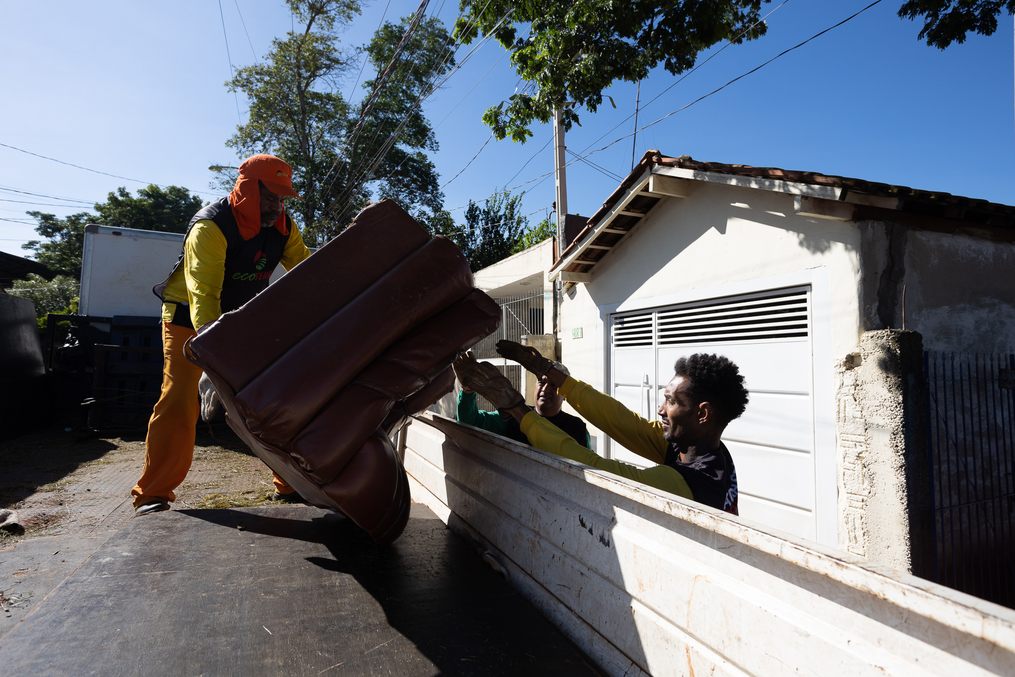 A imagem mostra três homens carregando um sofá antigo e pesado para dentro da carroceria de um caminhão. O homem da frente, vestindo uma camisa amarela e calças laranja, parece ser o principal responsável por carregar o sofá, segurando-o com firmeza. Ele usa luvas e um chapéu de proteção. Dois outros homens ajudam a levantar e posicionar o sofá na parte de trás do veículo, também usando luvas. Eles parecem estar colaborando de forma eficiente. O caminhão está estacionado em frente a uma casa modesta, com um portão de garagem branco. O dia é ensolarado e o céu está azul claro. A cena parece retratar uma atividade de coleta de lixo ou reciclagem de móveis. A imagem sugere trabalho em equipe e esforço físico.