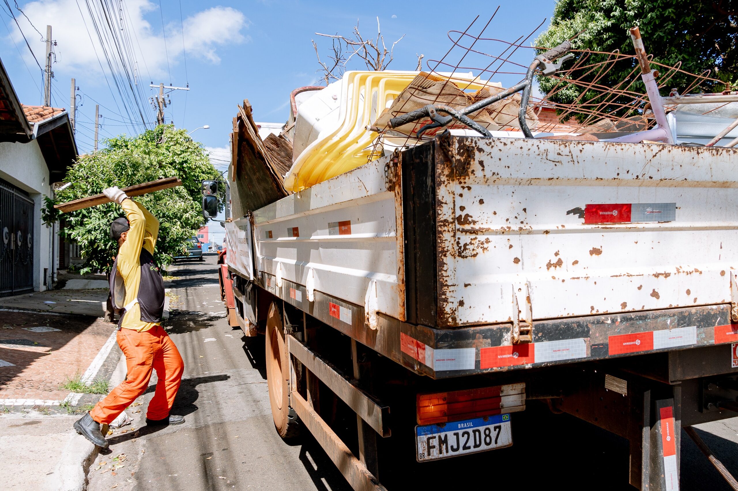 A imagem mostra um trabalhador de vestuário laranja e preto transportando um pedaço de madeira em direção a um caminhão de coleta de lixo. O caminhão está carregado com diversos itens, incluindo móveis e materiais metálicos, e está estacionado em uma rua urbana, cercada por árvores e casas ao fundo. A cena sugere um trabalho de limpeza ou remoção de entulho.