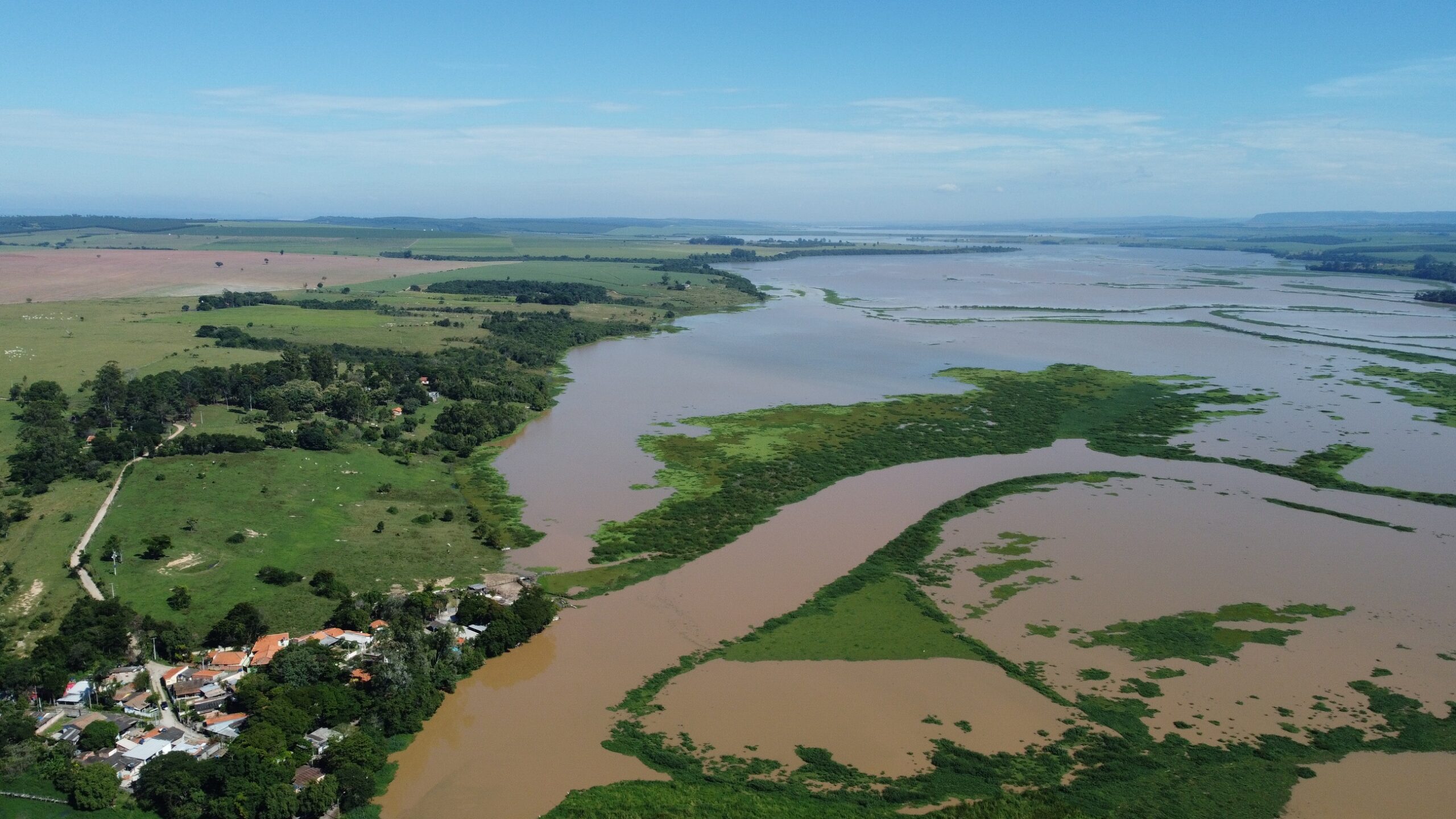 A imagem mostra uma vista aérea de uma paisagem onde um rio largo e sinuoso se encontra com uma área de terra verdejante. O rio parece ser largo e raso, com áreas de água mais escura e lama e áreas onde a vegetação emerge da água. Ao longo das margens do rio, há uma terra relativamente plana com pastagens e alguns agrupamentos de árvores. Uma pequena aldeia ou vila está visível perto da margem do rio, com edifícios em tons de terracota e rodeada de vegetação verde. A vegetação é composta por pastagens, pequenos grupos de árvores e, próximo à margem do rio, vegetação ribeirinha mais densa. No fundo da imagem, há uma paisagem mais plana e seca que parece ser campo cultivado. O céu está limpo e azul, com apenas algumas nuvens esparsas. A imagem transmite uma sensação de tranquilidade e calma, com um contraste harmonioso entre a água, a terra e o céu.