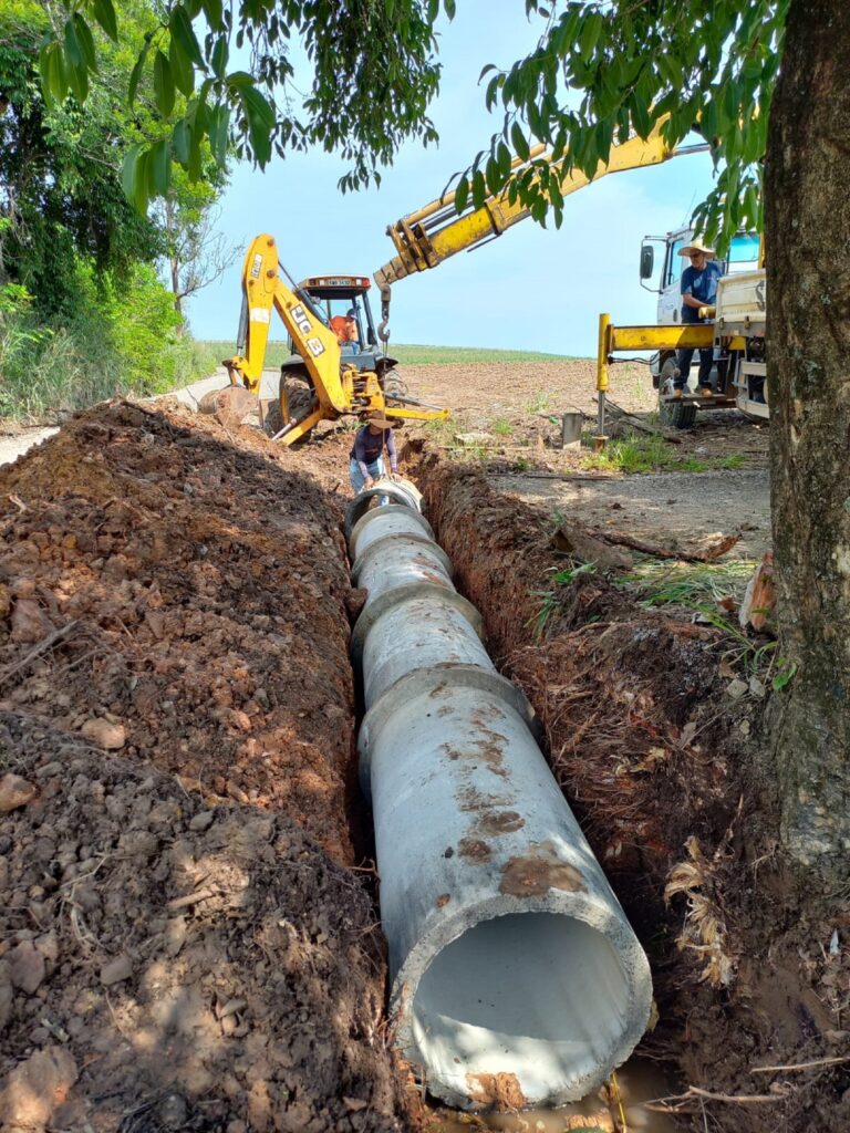 Tubo de galerias de águas pluviais sendo enterrado em uma vala, tendo a cima um homemn de chapéu empurrando.