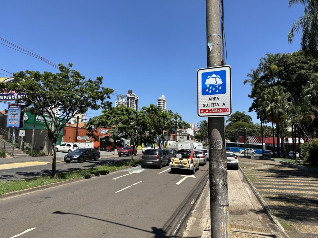 Imagem da avenida, mostra diversos carros nos dois sentidos, várias árvores e estabelecimentos comerciais. Em destaque, no canto direito, a placa instalada com os dizeres: ÁREA SUJEITA A ALAGAMENTO e o desenho de nuvens com gotas de chuva