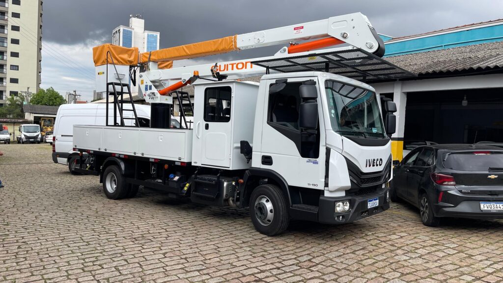 Caminhão branco com cesto duplo estacionado em pátio de paralelepípedo.