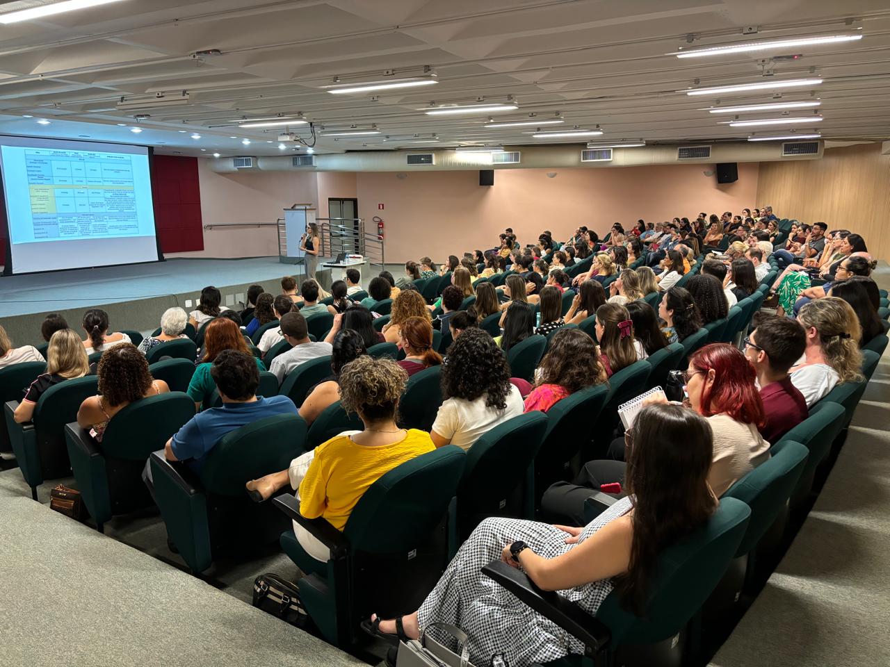 A imagem mostra uma palestra em um auditório. Uma palestrante está no palco, de frente para uma plateia sentada em cadeiras fixas em fileiras. Um projetor mostra um slide com informações sobre o uso de metformina, incluindo a necessidade de monitorar os níveis de vitamina B12 após quatro anos de uso devido ao risco de deficiência. O auditório é moderno, com iluminação artificial e paredes de cor bege. A plateia é composta por muitas pessoas, principalmente mulheres, que parecem estar atentas à palestra. A atmosfera é calma e profissional.