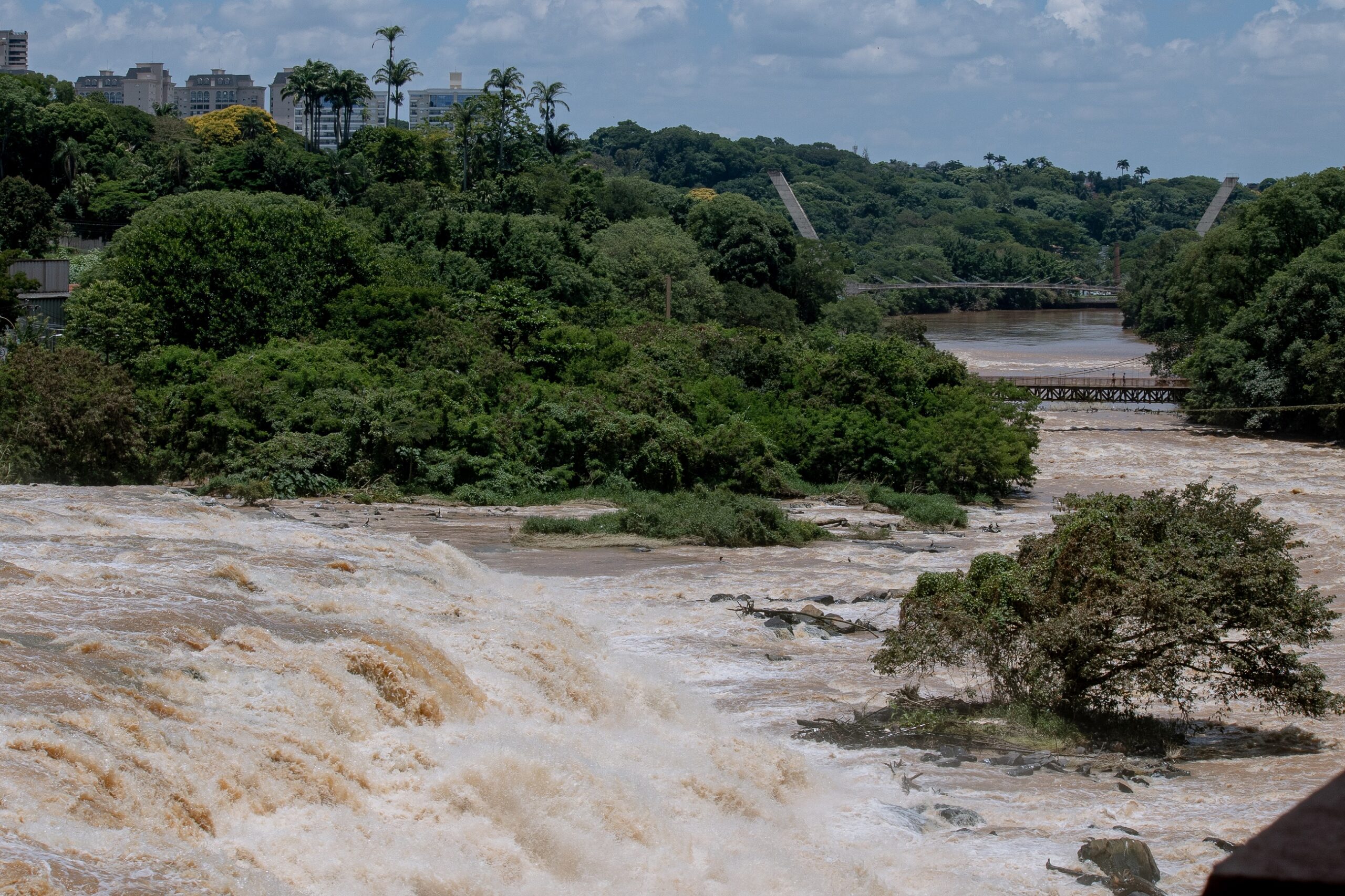 A imagem mostra uma cachoeira volumosa e turva com água marrom-esbranquiçada, correndo sobre rochas e se espalhando por uma área ampla. A água está agitada e espumosa, indicando um fluxo forte. Na parte de trás, há uma vegetação exuberante e verde, densamente florestada, que se estende até o horizonte. Podemos ver alguns edifícios de apartamentos no fundo à esquerda, indicando uma área urbana próxima. Uma ponte suspensa atravessa um rio mais calmo além da cachoeira, conectando as duas margens da floresta. Uma pequena ilha com uma árvore se encontra no meio do rio, logo abaixo da cachoeira. O céu é parcialmente nublado, com algumas nuvens brancas e algumas áreas de céu azul visíveis. O tom geral da imagem é natural e indica um dia ensolarado. A cena sugere uma área de natureza exuberante, com uma combinação de ambiente urbano próximo e a força da natureza.