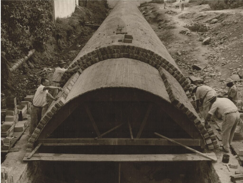 Imagem antiga mostra a canalização sendo feita, na década de 50; há cinco homens trabalhando, trajando calças claras e camisas brancas