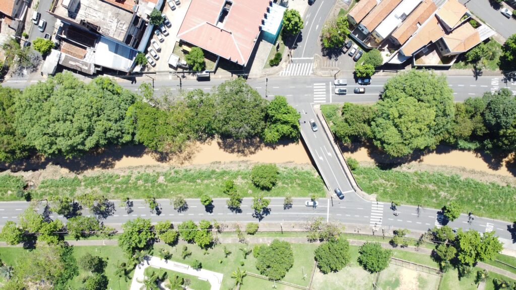 Imagem aérea do ribeirão; mostra muitas árvores no entorno e algumas casas ao fundo. Há avenida dos dois lados, com carros passando