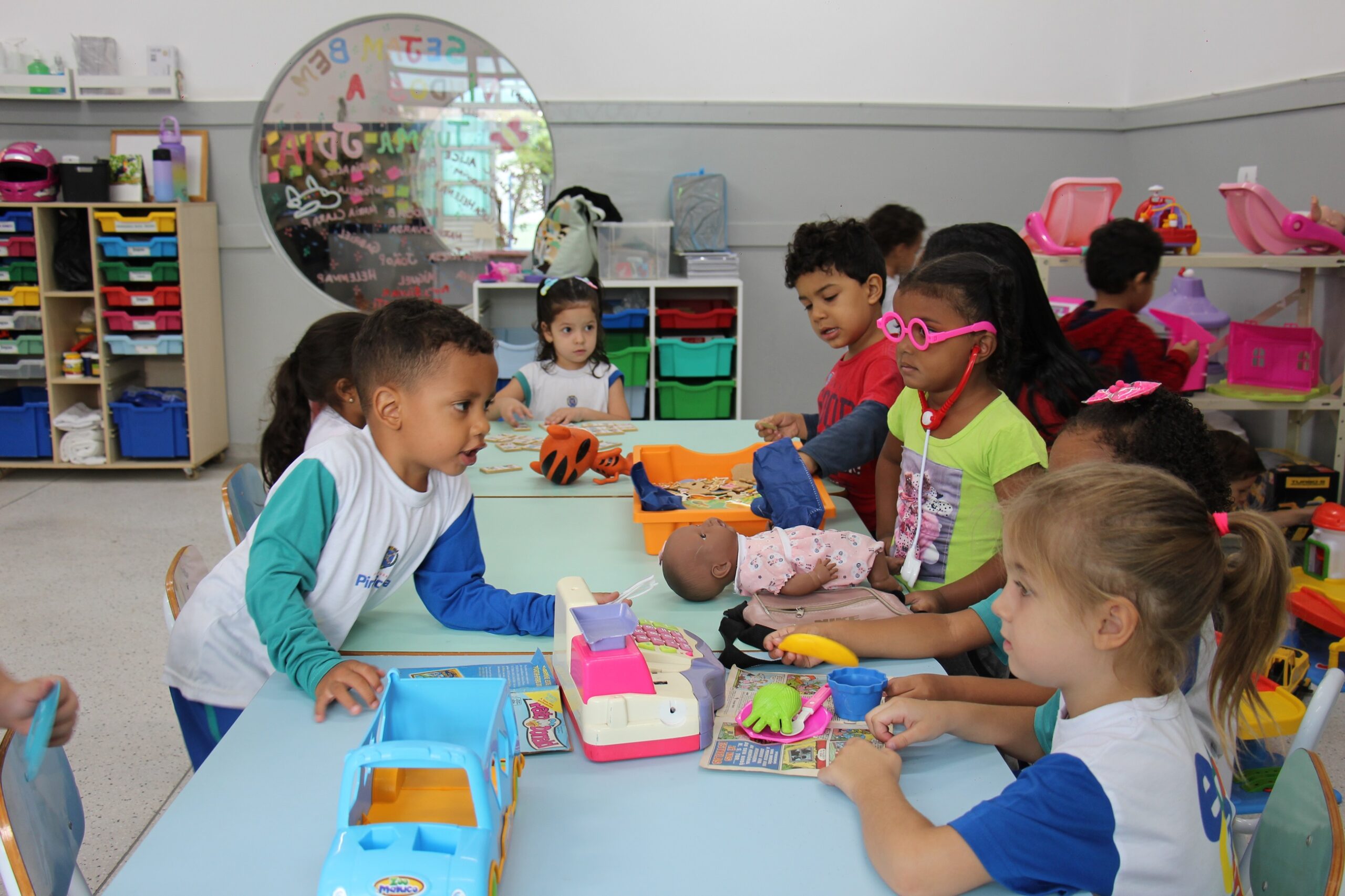 Descrição da imagem: Crianças em uma sala de aula brincando. A maioria das crianças estão sentadas ao redor de uma mesa azul clara, onde há vários brinquedos, como um carrinho, um telefone, uma boneca e outros objetos. As crianças estão interagindo entre si e com os brinquedos. Há prateleiras com caixas coloridas ao fundo e um espelho redondo na parede. A sala é bem iluminada e parece ser um ambiente de aprendizado e brincadeira. A criança mais próxima da câmera é uma menina loira com um rabo de cavalo, vestindo uma camiseta azul e branca. Ela está focada em algo na mesa. À esquerda dela, uma criança negra de camiseta verde e óculos cor de rosa brinca com uma banana de brinquedo. No centro, um menino negro de camiseta branca e verde interage com os brinquedos na mesa. Ao fundo, outras crianças brincam, e uma delas usa uma camiseta vermelha. Há uma variedade de brinquedos espalhados pela sala.