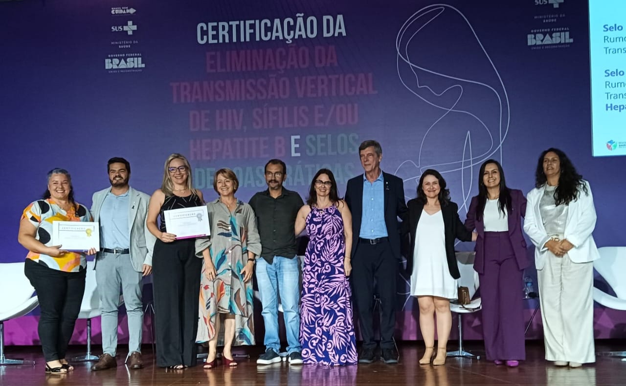 Esta imagem mostra uma cerimônia de certificação da eliminação da transmissão vertical de HIV, sífilis e/ou hepatite B no Brasil. Várias pessoas, incluindo autoridades e profissionais de saúde, estão presentes e segurando certificados. Há um banner atrás deles com a informação sobre a certificação e os selos do programa. A imagem captura um momento de celebração e reconhecimento pelo sucesso do programa de saúde pública.