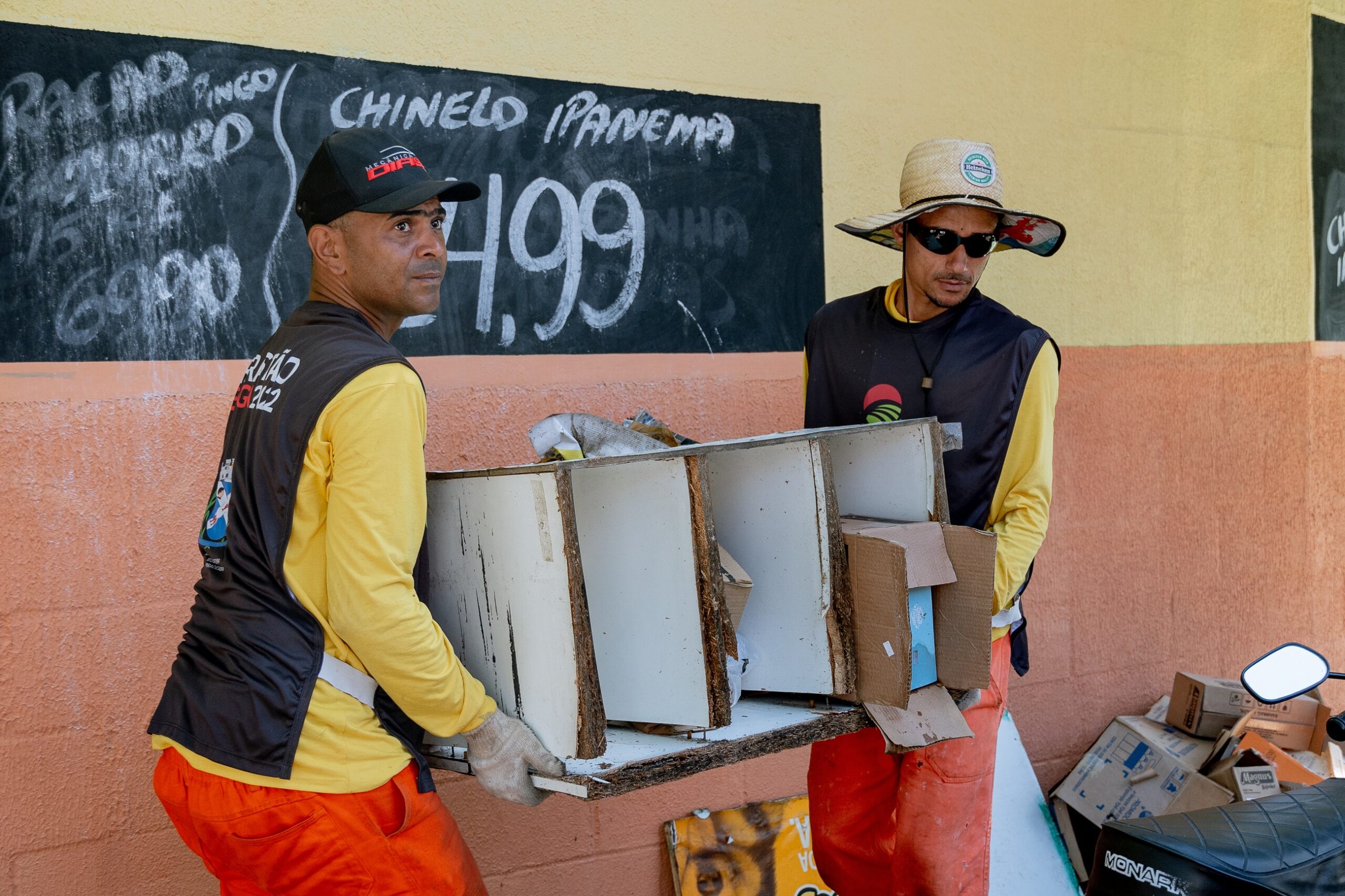 A imagem mostra dois homens vestindo uniformes de trabalho, carregando juntos um pedaço grande de mobília de madeira velha e danificada. A mobília parece ser um pedaço de armário ou estante, e está em más condições. Os homens parecem estar trabalhando em uma tarefa de limpeza ou remoção de entulho. O homem à esquerda usa um boné de beisebol preto e uma camisa amarela de mangas compridas sobre um colete preto. Sua calça é laranja. O homem à direita usa um chapéu de palha largo e óculos de sol, com uma camisa amarela de mangas compridas por cima de um colete preto. Ele também usa calças laranja. Eles estão em frente a uma parede de cor creme com uma lousa preta na qual está escrita, em letras brancas, uma propaganda ou preços de algum produto. Uma moto parcialmente visível está no canto inferior direito da imagem, em frente a uma pilha de caixas e outros objetos. O ambiente parece ser uma rua ou área externa de uma cidade, possivelmente perto de um negócio. A imagem tem uma iluminação natural, com sombras sutis. A cena transmite a ideia de trabalho árduo e esforço físico.
