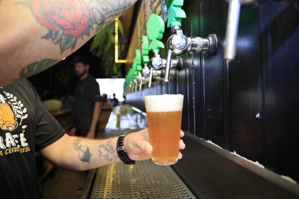 A imagem mostra um barman com tatuagens nos braços, servindo uma cerveja artesanal. A cerveja está sendo despejada de um torneira de chope em um copo curto e largo. A torneira é cromada e tem uma pequena placa verde acima dela, indicando possivelmente o tipo de cerveja. O barman usa uma camiseta escura com uma imagem de um caneco de cerveja e o que parece ser o nome de uma cervejaria. Ao fundo, um homem de camisa escura está ligeiramente fora de foco, mostrando que a atenção principal está no ato de servir a cerveja. A área do bar é moderna e limpa, com um painel preto atrás das torneiras. A iluminação é suave e cria uma atmosfera aconchegante. A imagem transmite uma sensação de qualidade e cuidado, tanto na cerveja como no ambiente do bar.