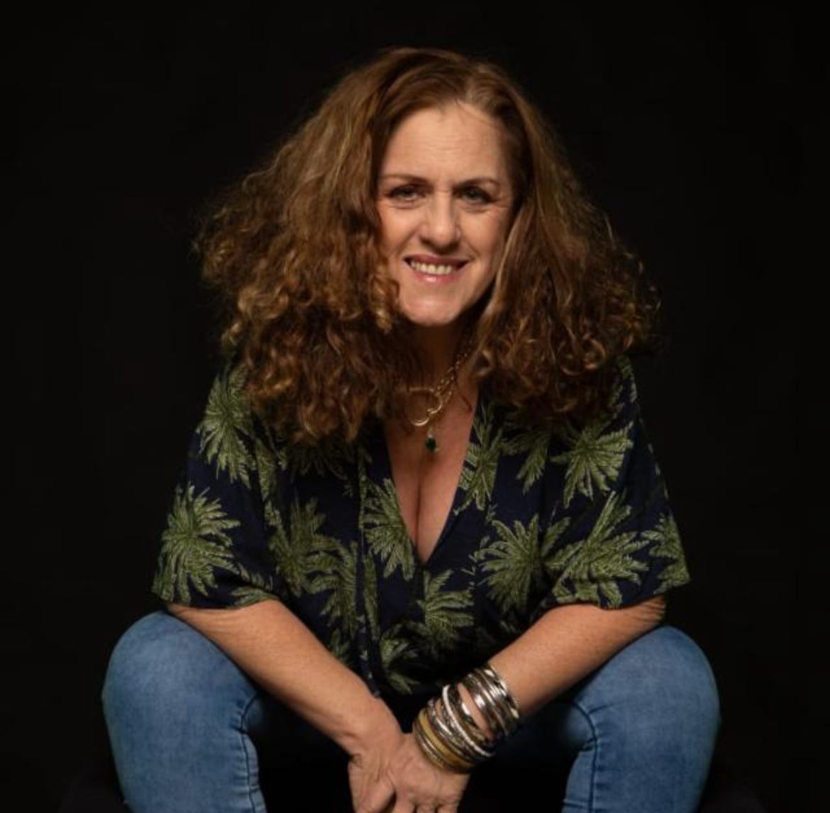 Mulher aparece sentada vestida de calça jeans e camisa verde e preta, sorrindo