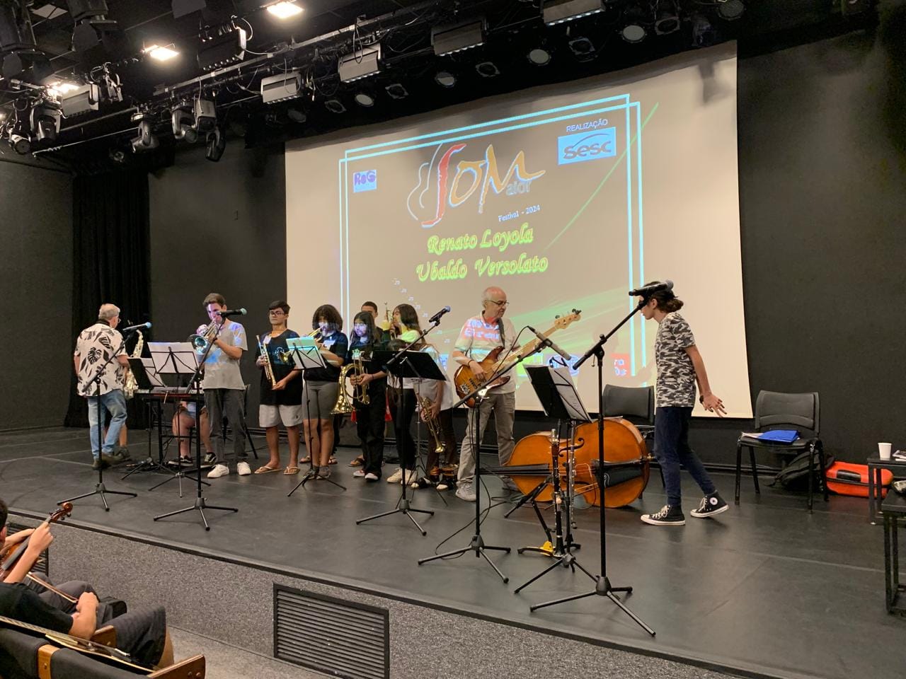 Crianças e adolescentes estão em um palco, em um ensaio, com instrumentos musicais