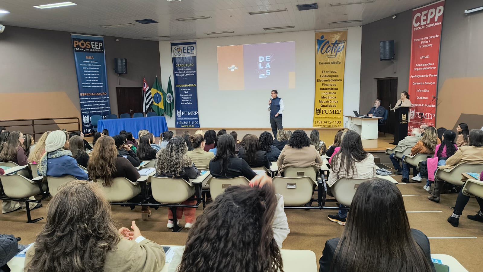A imagem mostra uma palestra em uma grande sala de aula. O palestrante está no palco, apresentando para uma plateia numerosa de pessoas sentadas em cadeiras organizadas em fileiras. Há dois painéis grandes na parede atrás do palestrante com informações sobre cursos e programas educacionais, em português. Há bandeiras brasileiras à esquerda da imagem. As pessoas na plateia são em sua maioria mulheres, e estão vestindo roupas casuais. O ambiente parece ser uma universidade ou escola. A iluminação é artificial, típica de uma sala de aula interna. A imagem é bem iluminada e focada. Há cartazes na sala com os logotipos da "FUMEP" e "CEPP", sugerindo uma instituição de ensino.