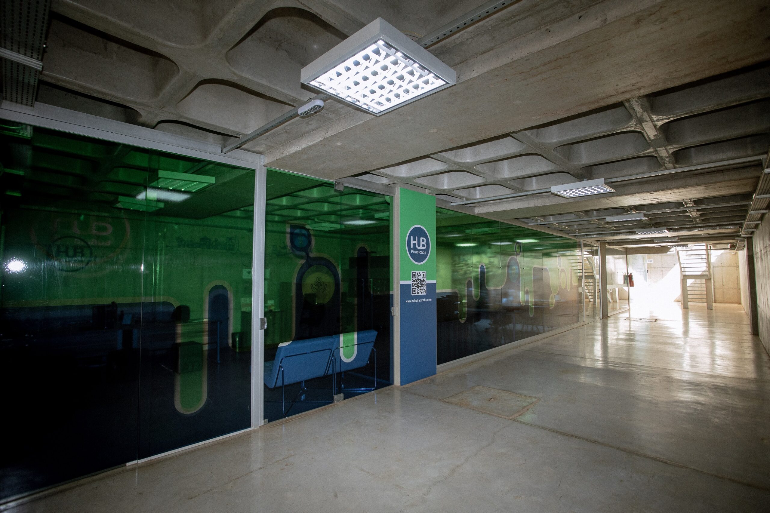 A imagem mostra um corredor longo e estreito com paredes de vidro de um lado e uma parede de concreto do outro. O corredor é iluminado por luzes fluorescentes no teto. Há uma porta visível no final do corredor. As paredes de vidro mostram um design verde e abstrato. Há um letreiro com a sigla "HB" visível em uma das paredes de vidro. Há uma pequena sala com um sofá azul atrás das paredes de vidro. O chão é de concreto polido. A imagem parece ser de um escritório ou de um espaço público. O ambiente parece limpo e organizado.
