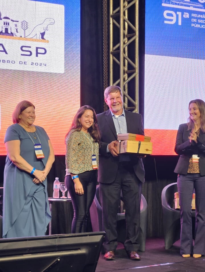 Imagem da titular da semuttrabn, Jane Franco, recebendo a homenagem das mãos de um home vestido com blazer e calça azuk